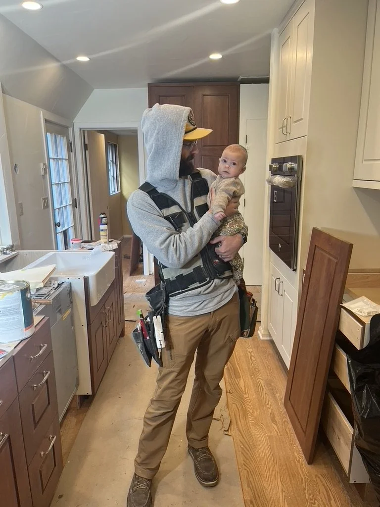 A man wearing a gray hoodie, tan pants, and a tool belt, holding a baby in a kitchen. The man has a beard and glasses, and the baby is dressed in a sweater and patterned pants, looking at the camera.