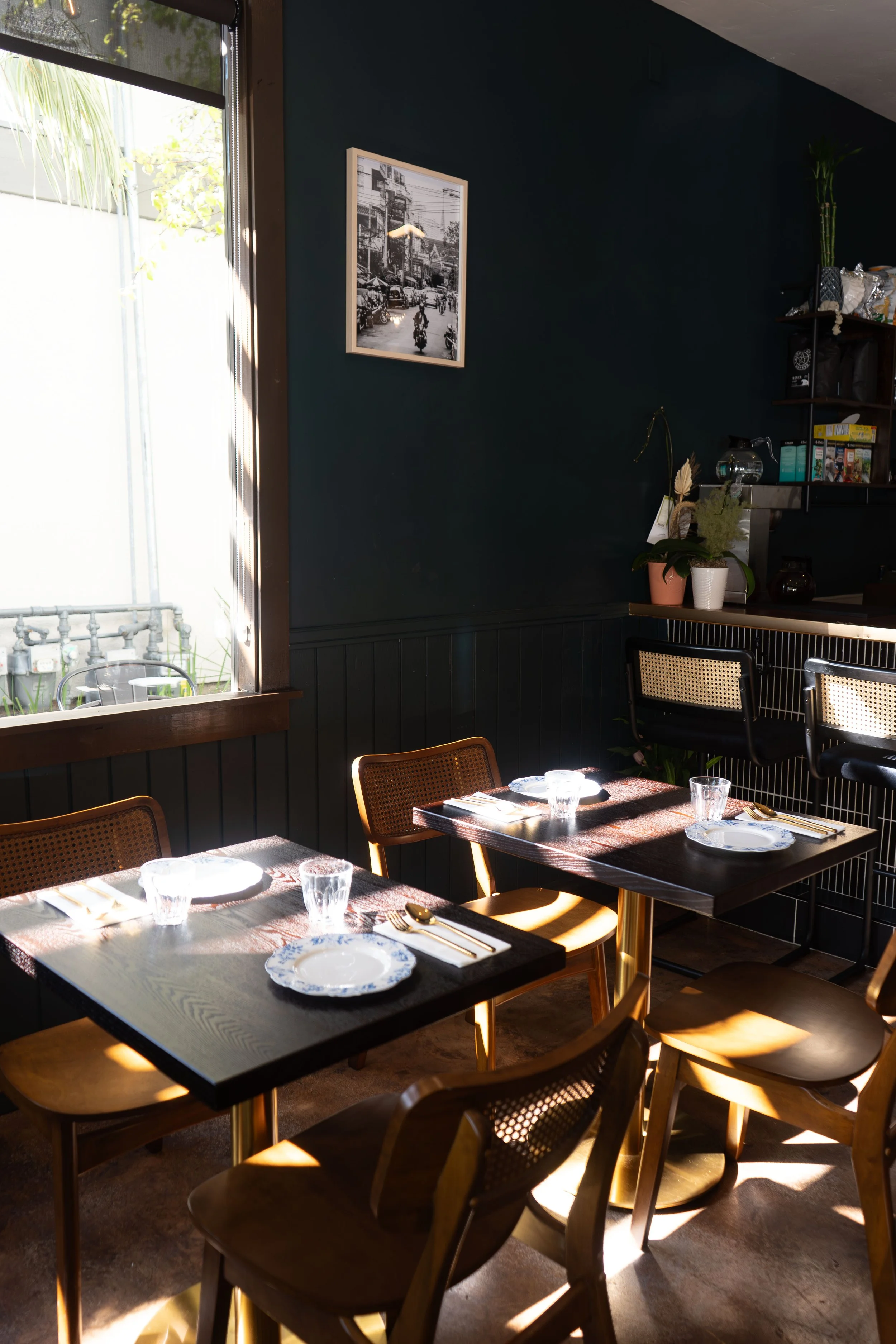 Interior dining room showing 2 2-top dark wood tables with gold utensils, white porcelain plates, clear water glasses, and modern rattan chairs. One wall features one Thai retro-themed framed photo of a city in Thailand, and a portion of coffee bar.