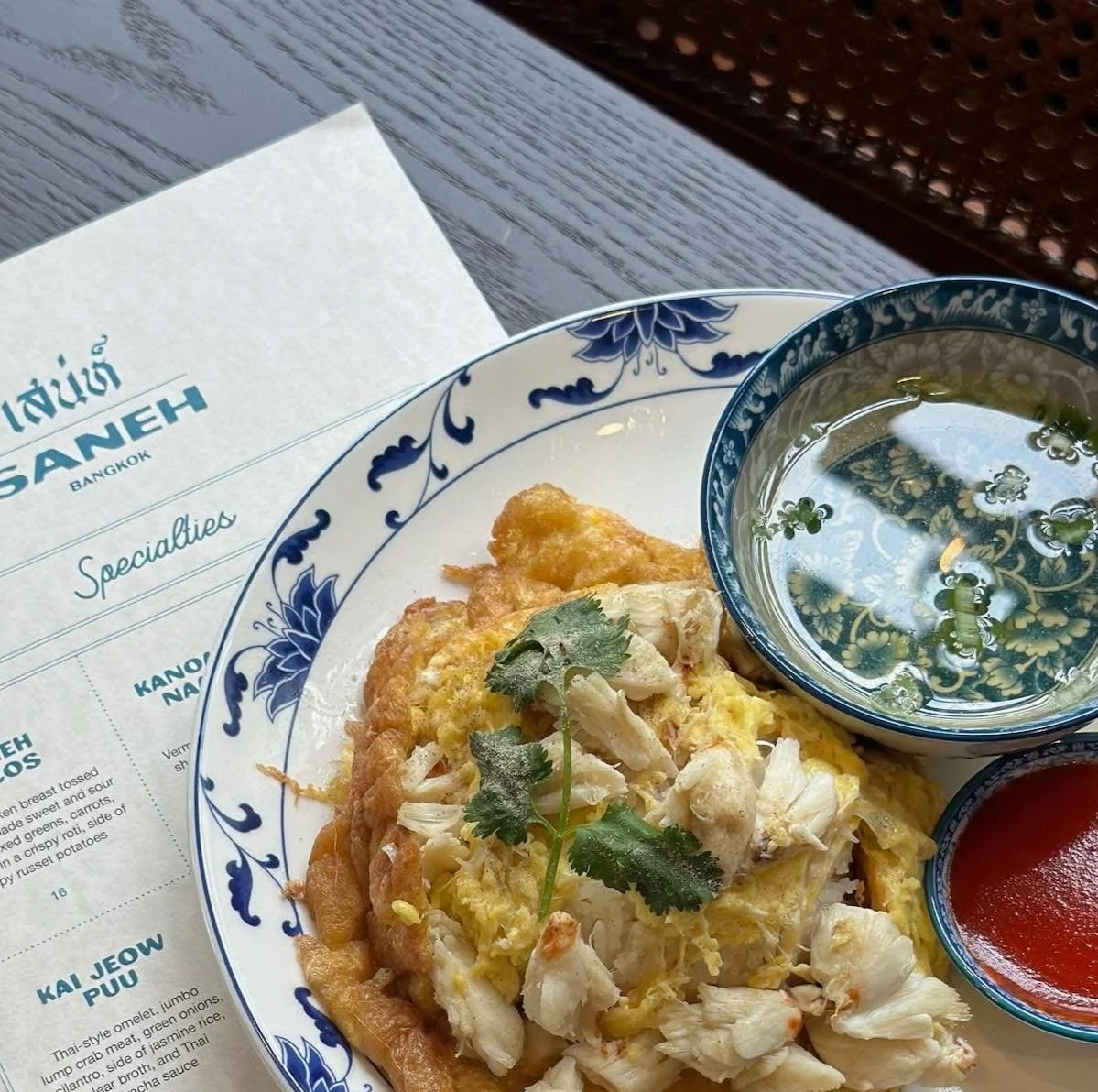 Thai-style crab omelet garnished with cilantro, side of clear broth in small bowl and Thai sriracha sauce, served on large white plate, and background of Saneh's specialties menu