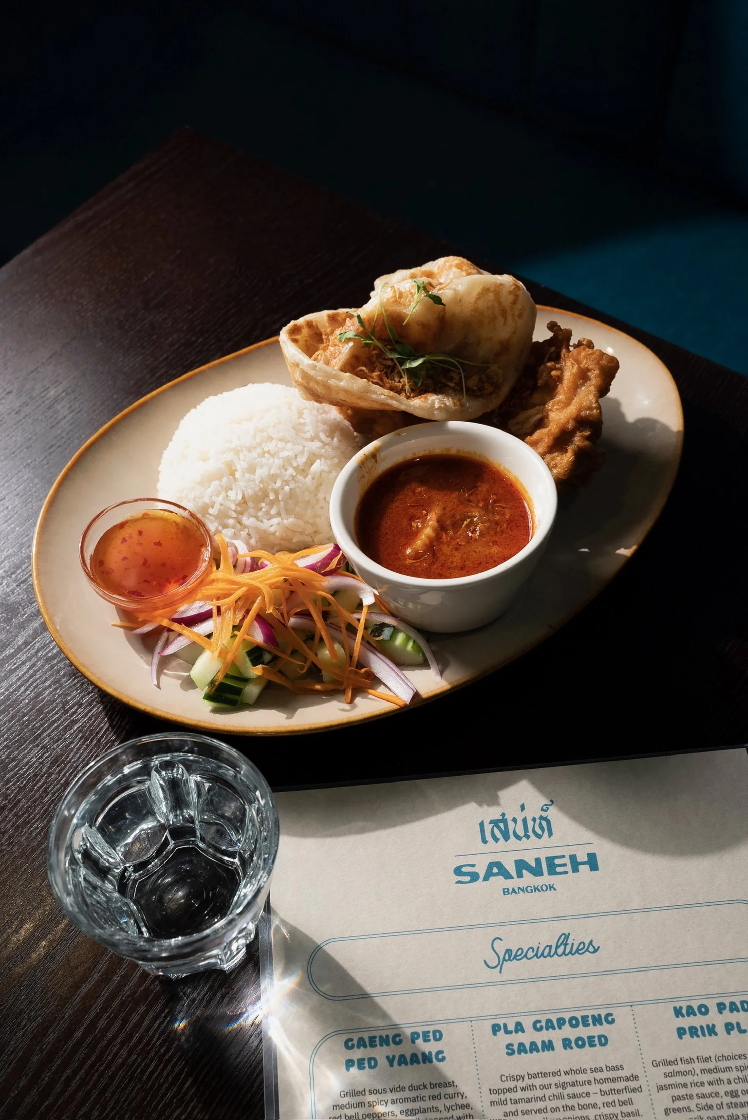Plate of brown fried chicken topped with one roti flatbread and microgreens, white jasmine rice, cucumber salad, and sweet and sour sauce, cup of water, and partial view of Saneh's Specialties menu