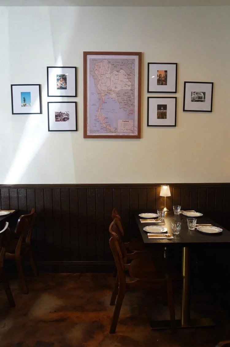 Interior dining room showing 1 dark wood table with gold utensils, white porcelain plates, clear water glasses, a table lamp, and modern rattan chairs. One wall features gallery of Thai retro-themed framed map of Thailand and photography.