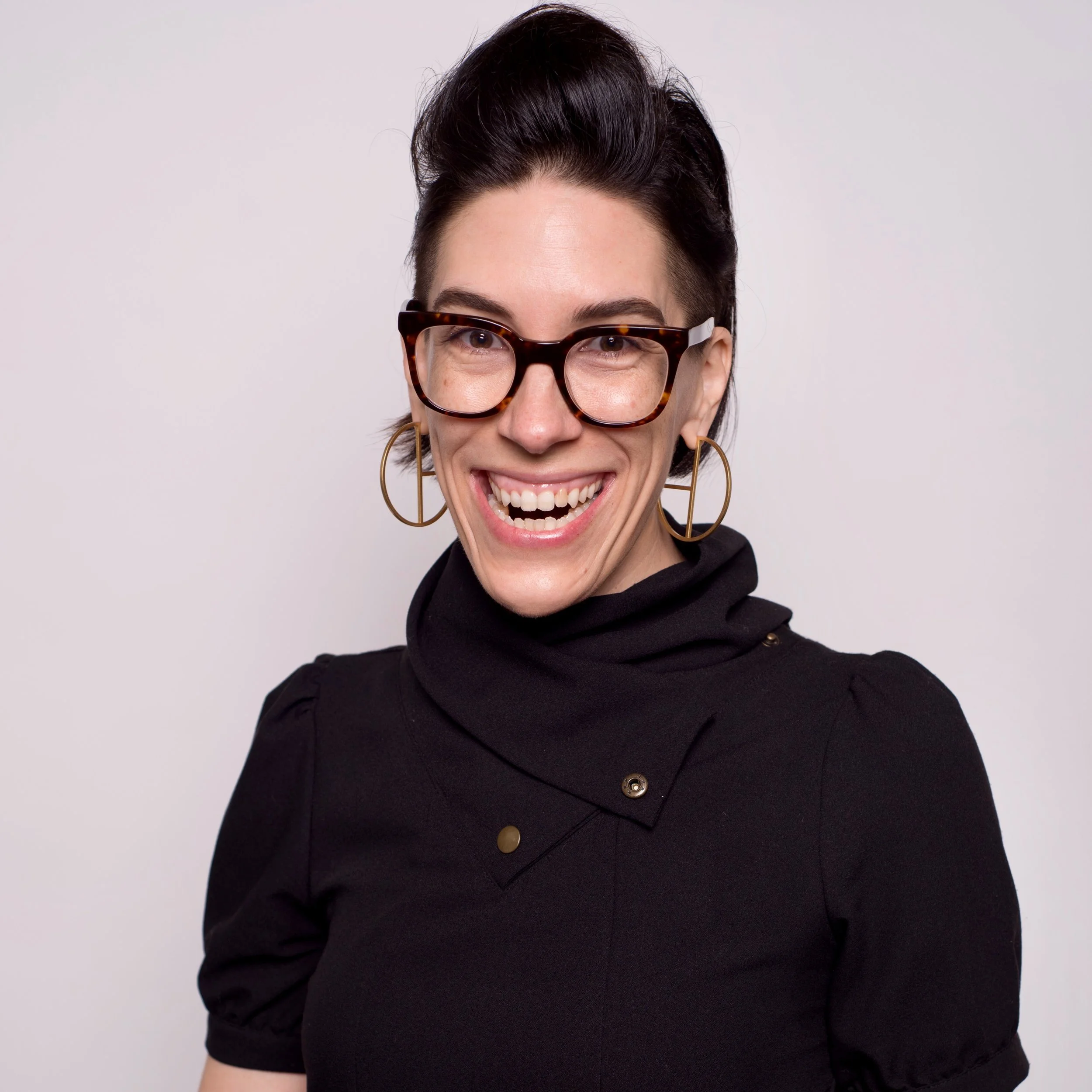 Woman smiling widely while wearing glasses and large hoop earrings, dressed in a black top with a high collar. She has dark hair styled up, and the background is plain and light-colored.