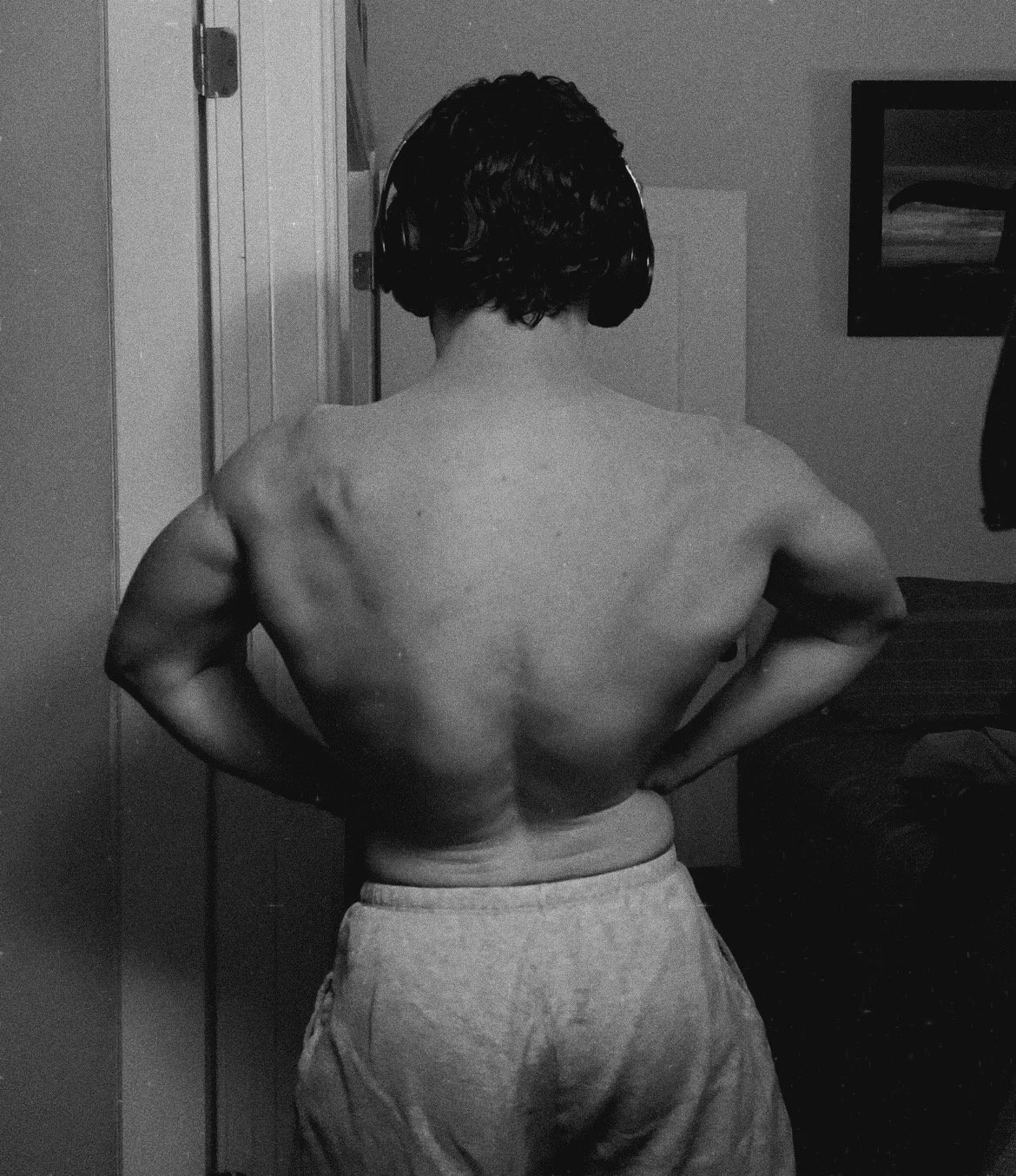 Back view of a shirtless man with headphones, standing with hands on hips in a room.