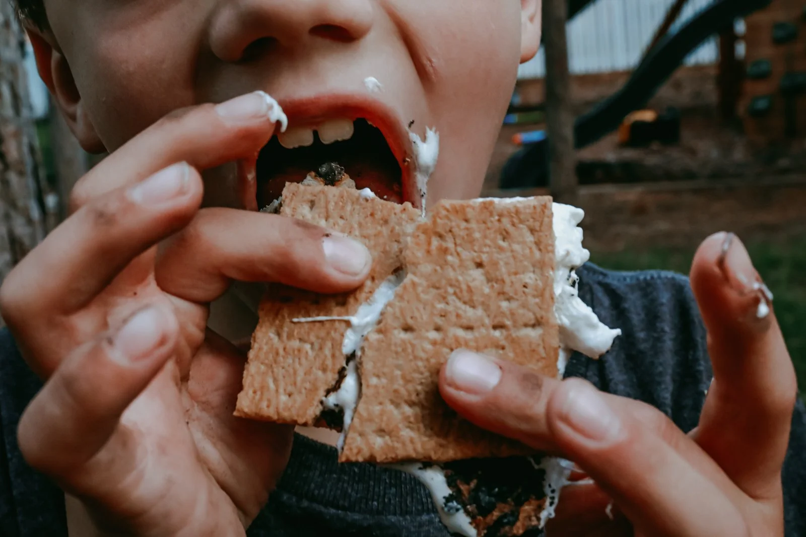 A child is eating a s'more with graham cracker, chocolate, marshmallow, and melted cream outside.