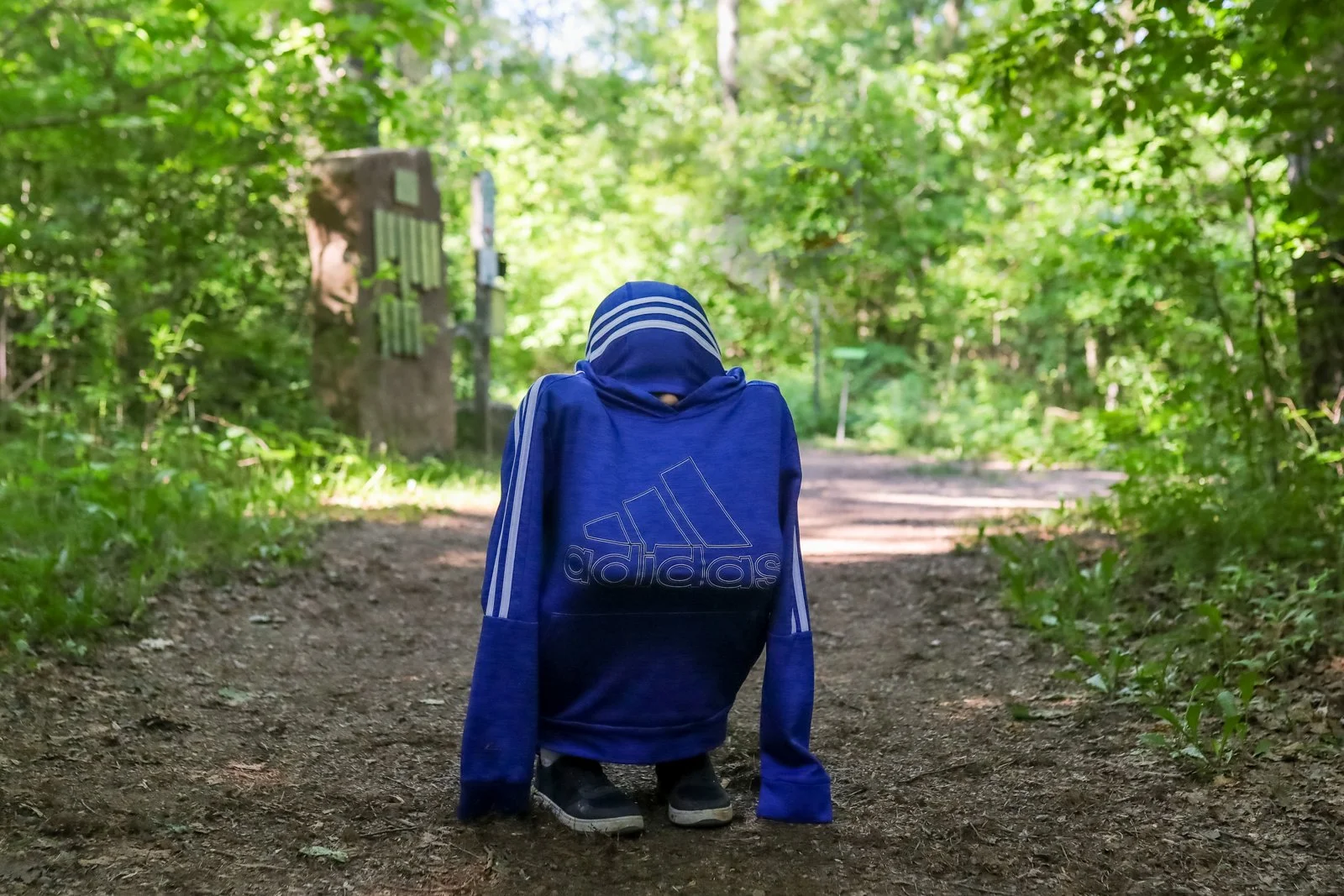 A child wearing a blue Adidas hoodie and shoes walks down a wooded dirt path with lush green trees surrounding the trail.