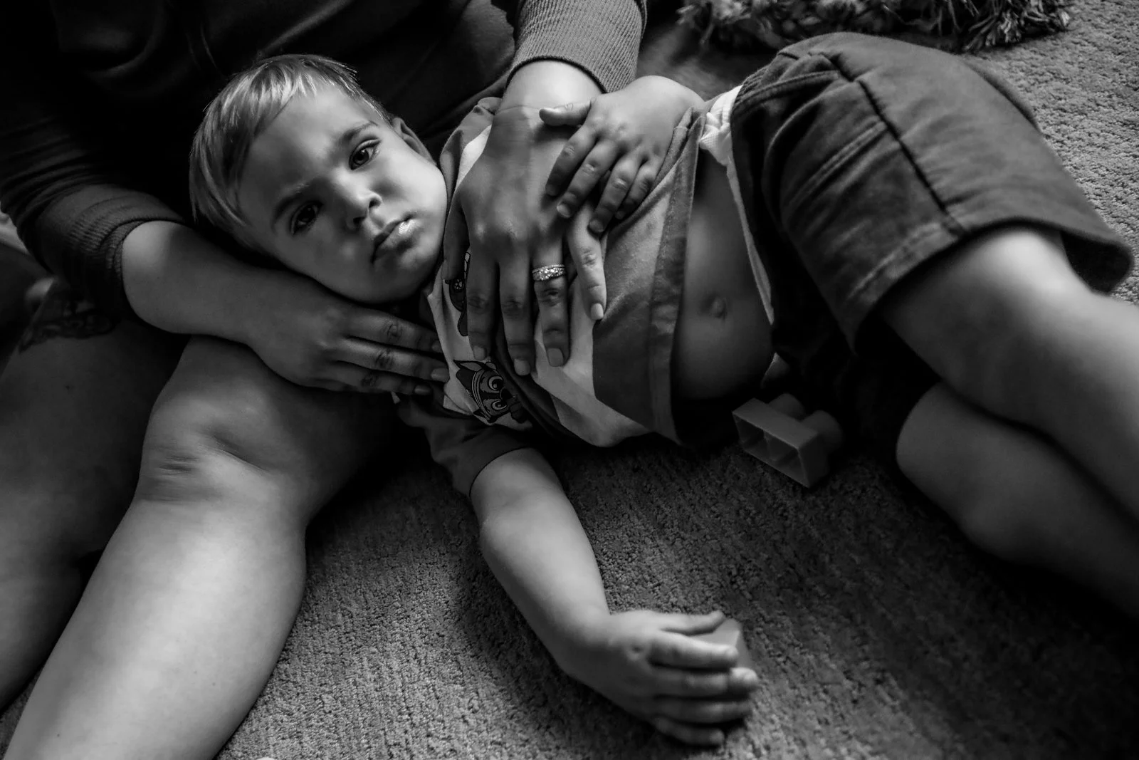 Child lying on the floor with a serious expression, surrounded by adults' hands touching them.
