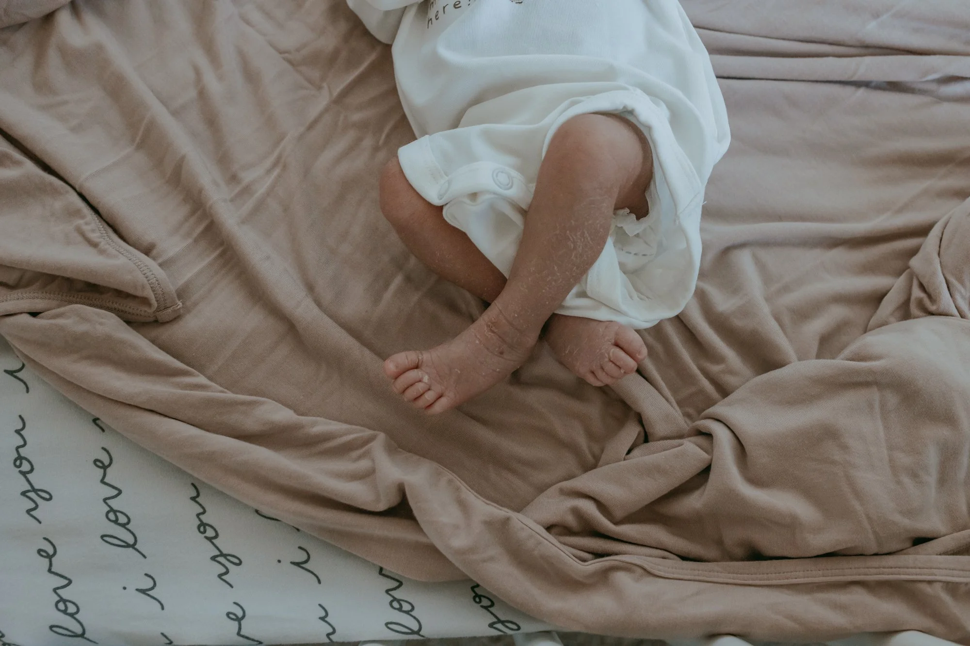 A baby lying on a bed, partially covered with a beige blanket, wearing a white onesie, with only legs and a portion of the arm visible.