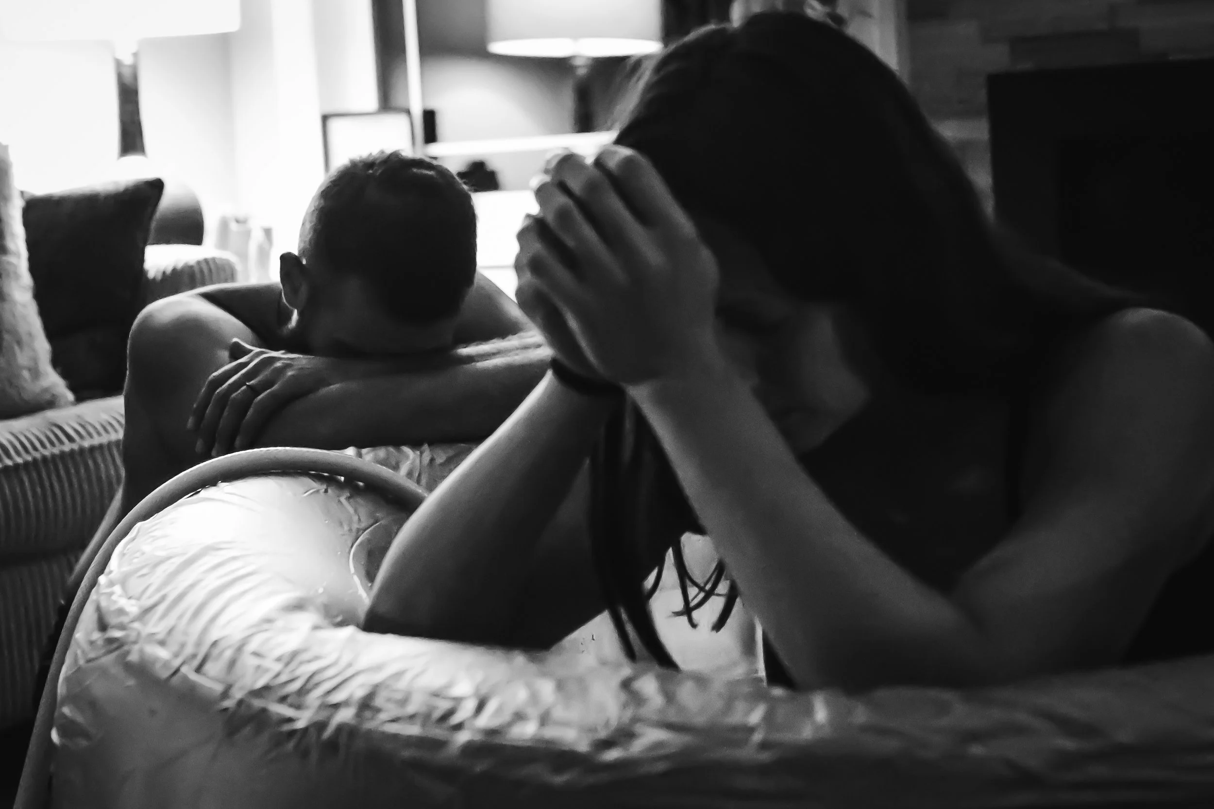 Black and white photo of a woman holding her head in her hands, sitting on a bed, with a young boy resting his head on a pillow in the background in a cozy living room.