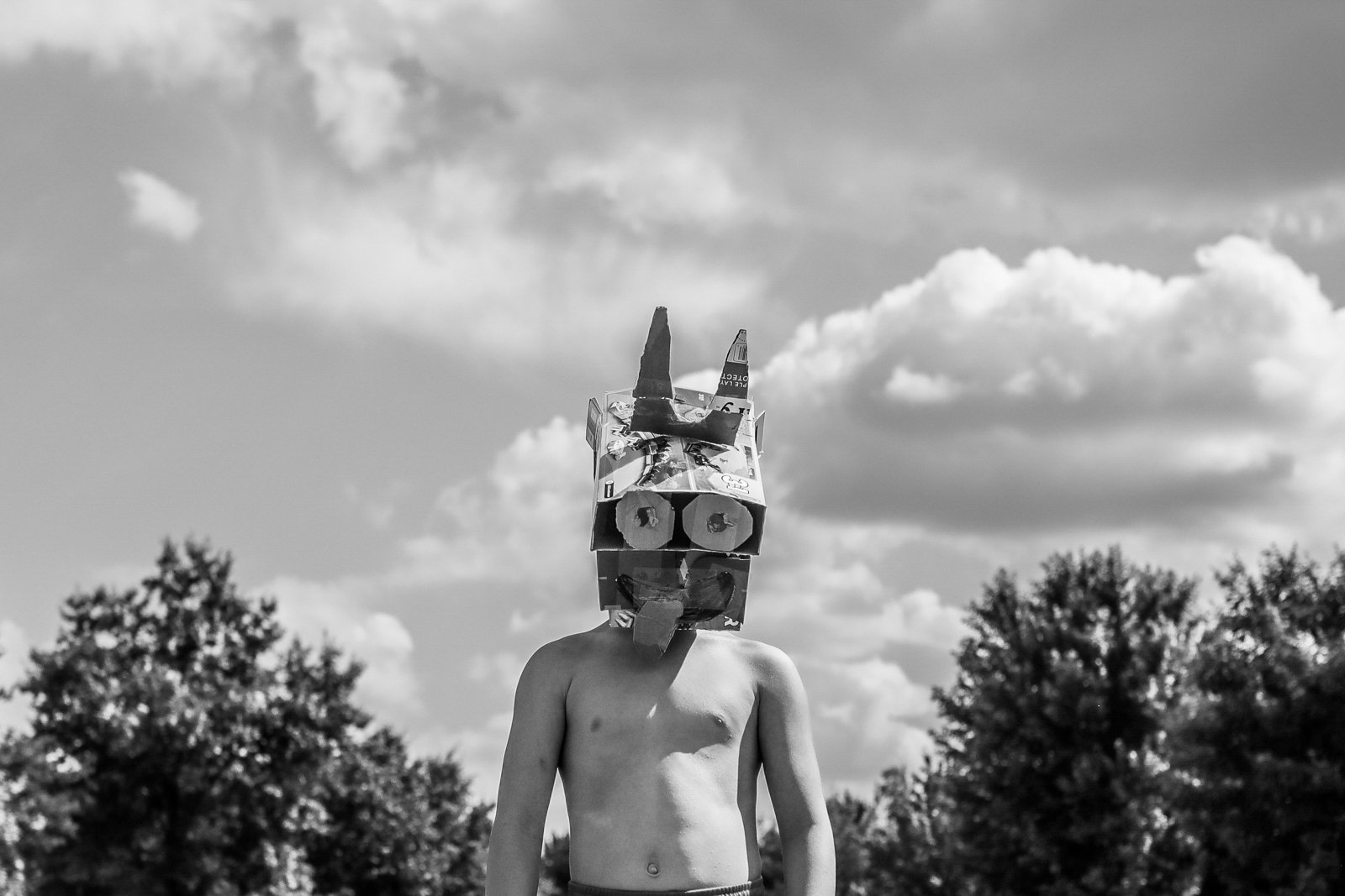 A shirtless boy wearing a cardboard robot mask standing outdoors with trees and clouds in the background.