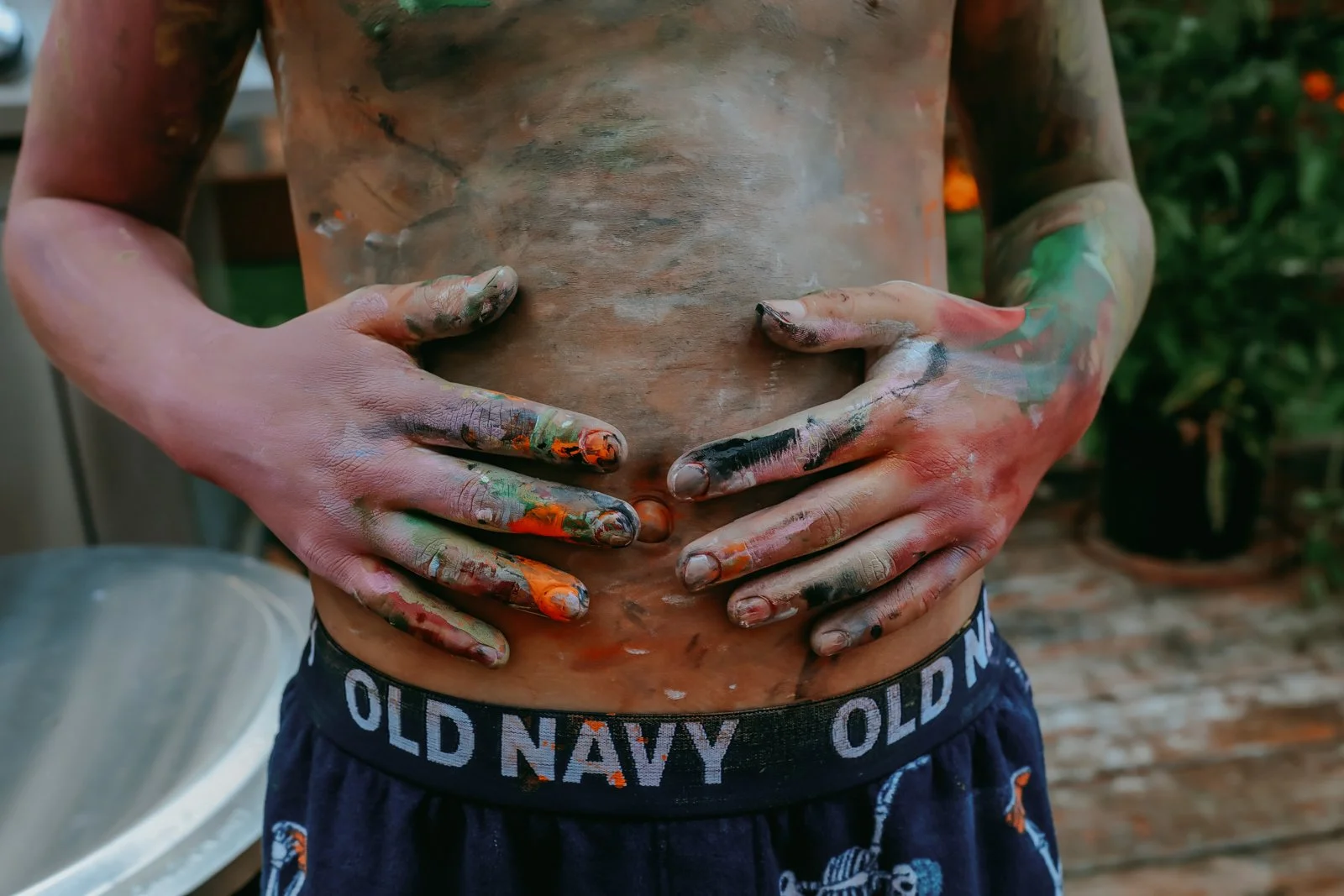 Person with hands covered in multicolored paint resting on their bare stomach, wearing navy Old Navy shorts, outdoors with greenery in the background.