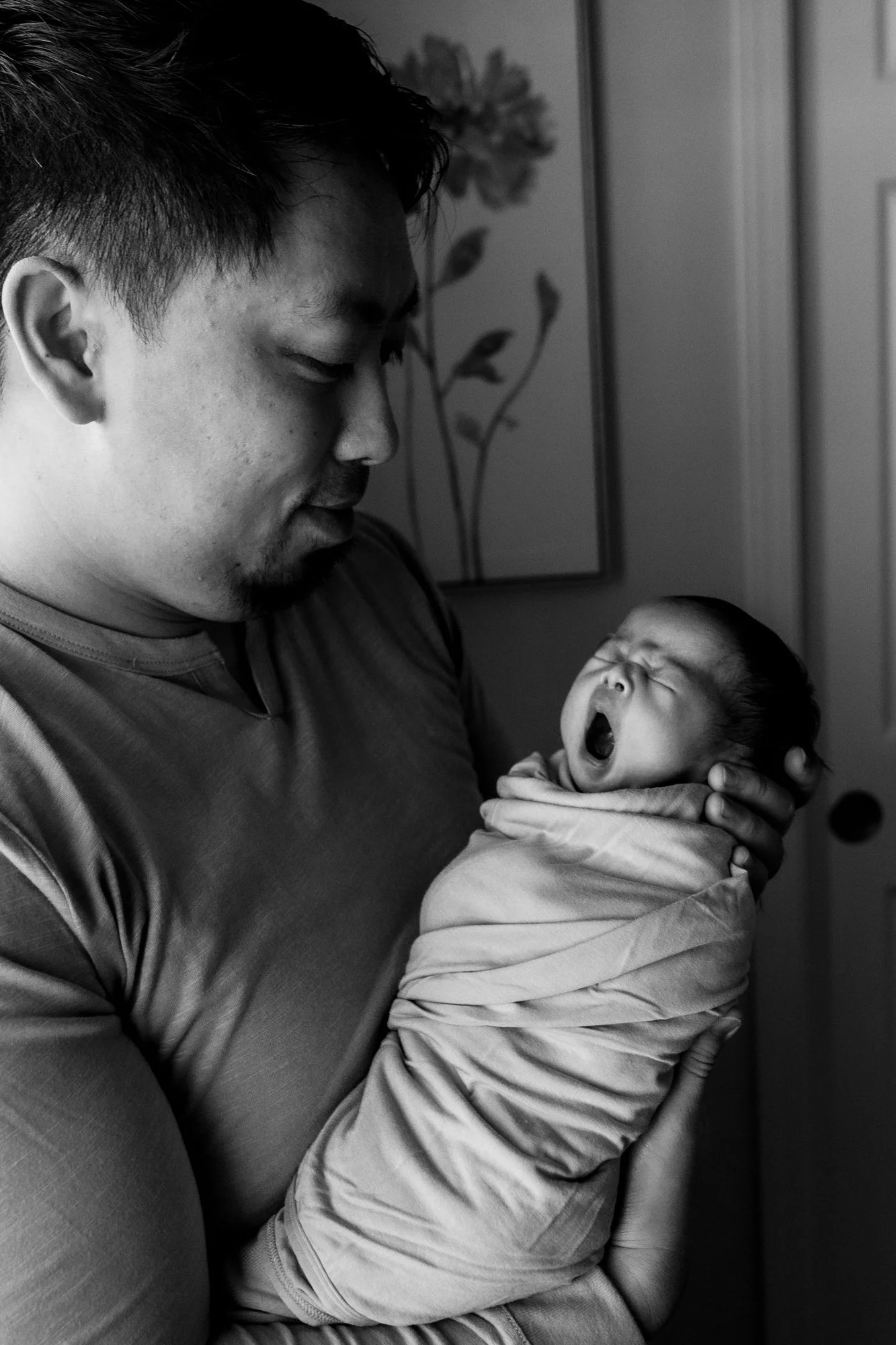 A man holding a yawning newborn wrapped in a blanket indoors.