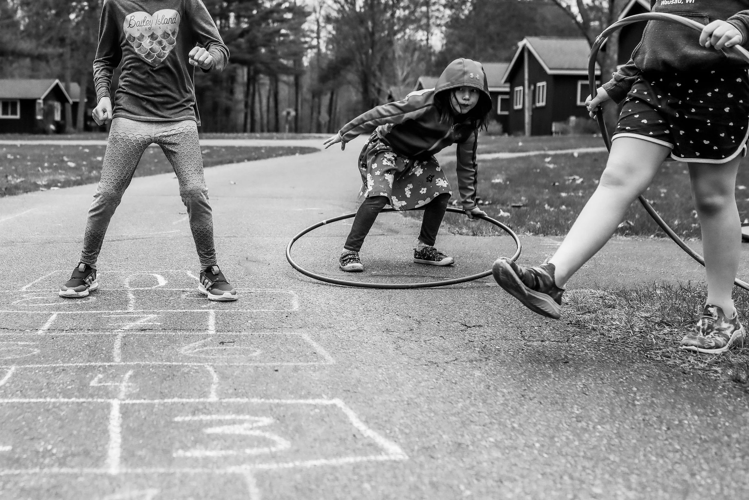 Children playing hopscotch and with hula hoops outdoors in a neighborhood park.