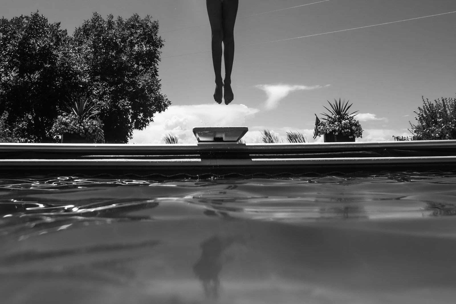Person diving into a swimming pool, viewed from the water level with trees and a partly cloudy sky in the background.