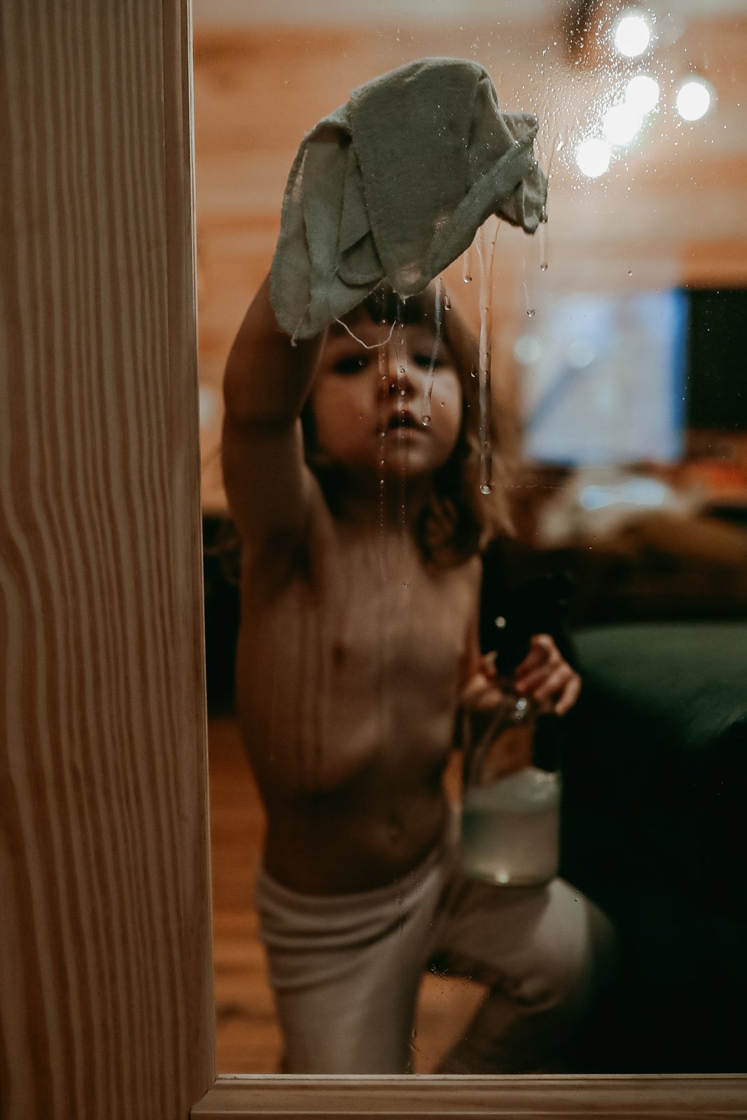 A young child with long hair takes a shower, holding a wet cloth up to a glass shower door, with water droplets visible on the glass.