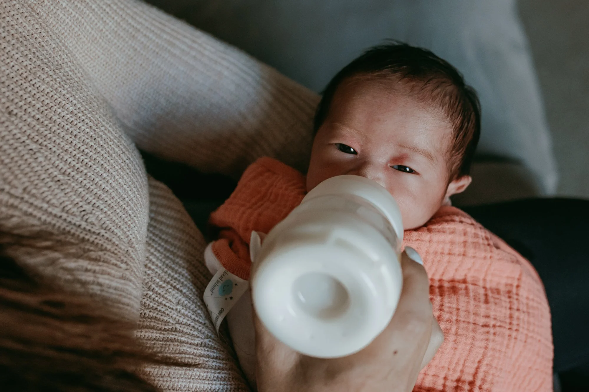 A baby with dark hair drinking from a baby bottle held by a person, lying on a beige couch with textured fabric, wearing an orange outfit.
