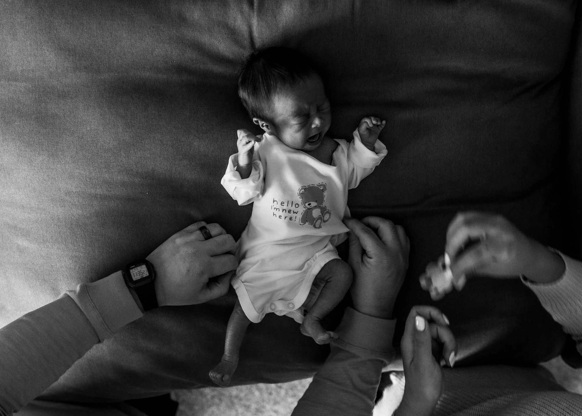 A crying newborn baby is lying on a couch, wearing a bib with a teddy bear graphic and the text 'hello! I'm new here!'; two adults are tending to the baby, one holding its hand and the other holding a small bottle.