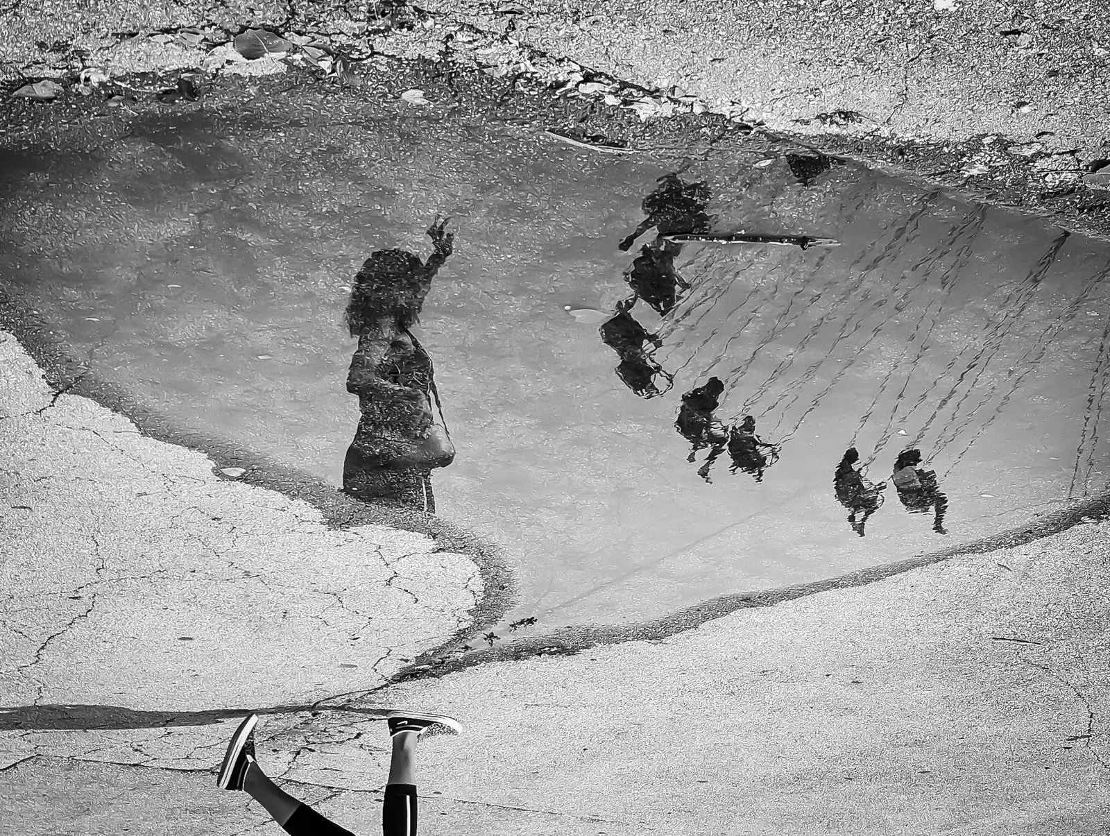 Reflection of children on a wet pavement, some on a swing set and others standing nearby, with a child lying on the ground in the foreground.