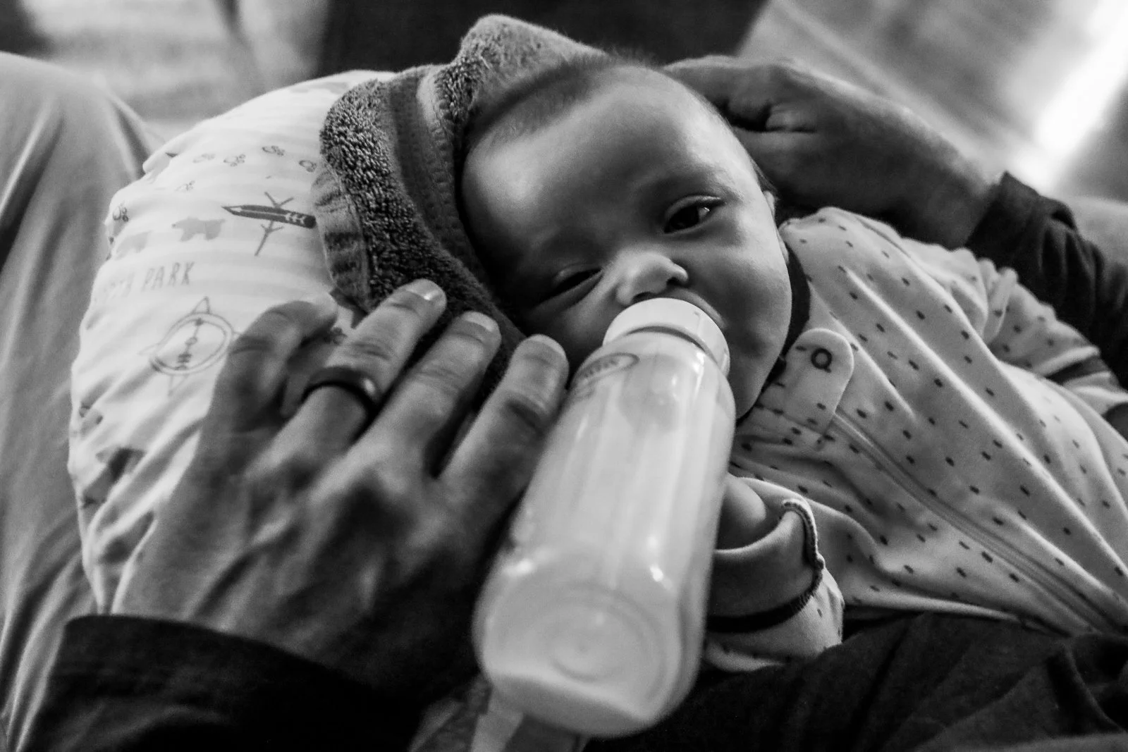A young child with a hooded jacket is lying on an adult's lap, drinking from a bottle, with the adult's hand gently touching the child's head.