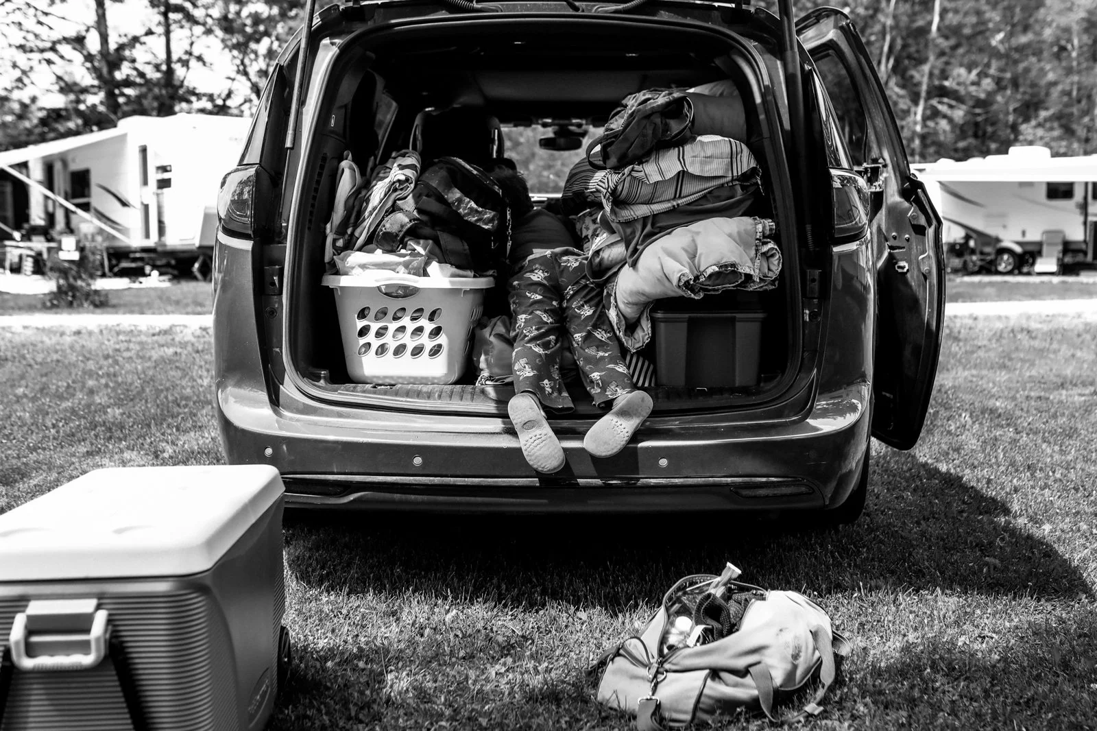 Open car trunk filled with clothes, bags, and household items, with a child's legs and shoes hanging out. Camping gear is placed on the grass outside the car.