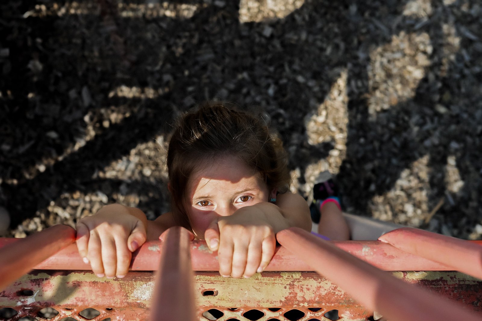 A young girl climbing a ladder, looking up at the camera with sunlight casting shadows across her face and arms.