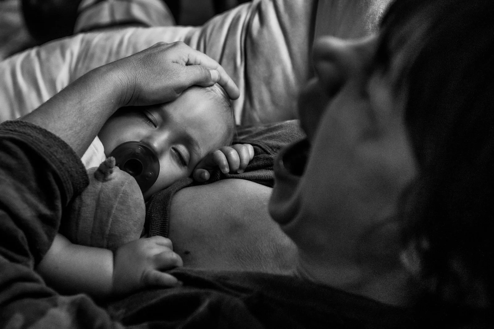 A young child with a pacifier resting on an adult's chest, held gently by an adult's hand, lying on a couch.