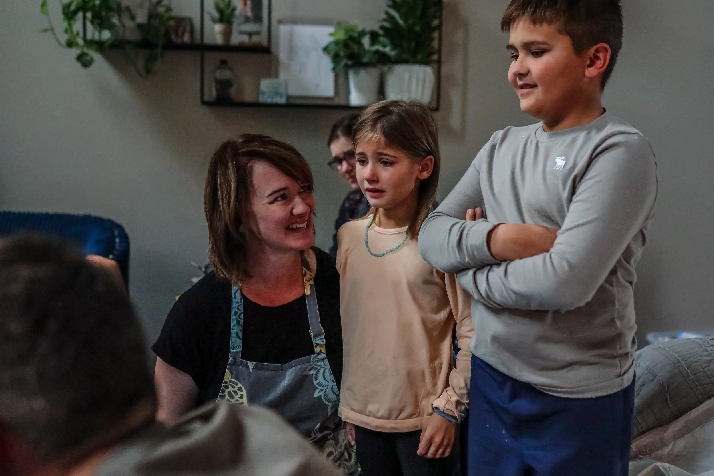 A woman in a black top and apron is talking to a boy with crossed arms and a girl looking worried or sad. Two other people are partially visible in the background in a room with shelves with plants and decorative items.