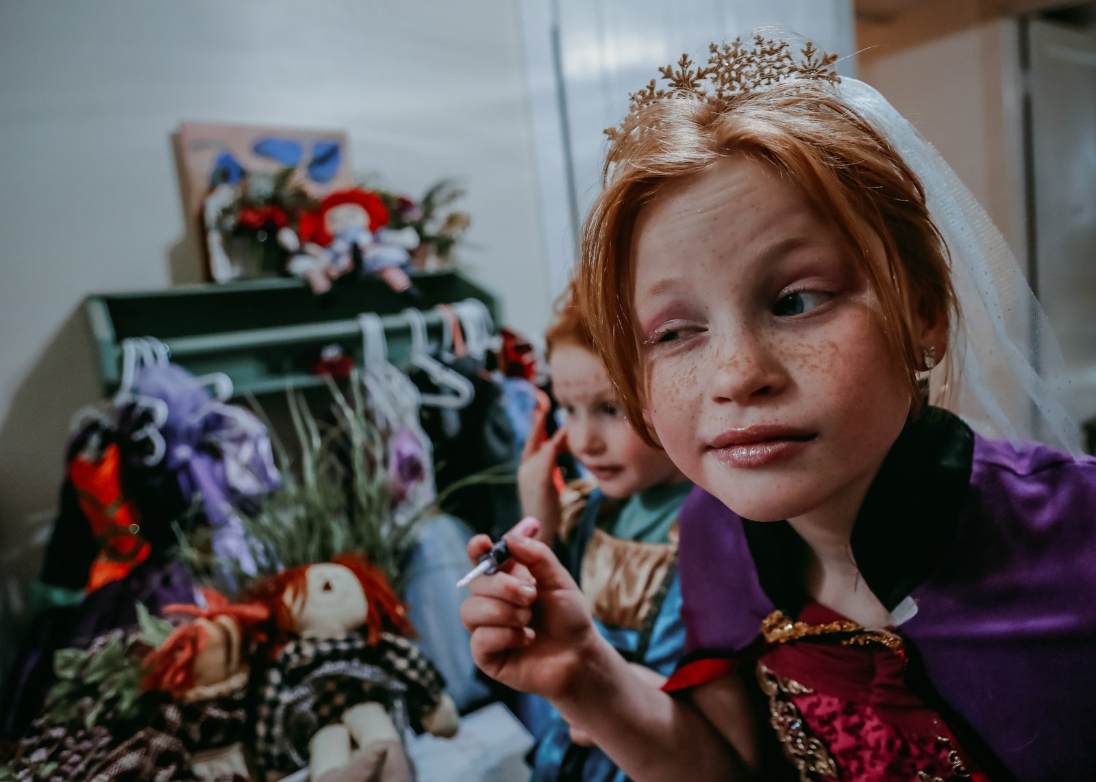 Two young children in costumes, one with a red wig and a purple cape, and the other with a green shirt, sitting with Halloween or holiday decorations in the background.