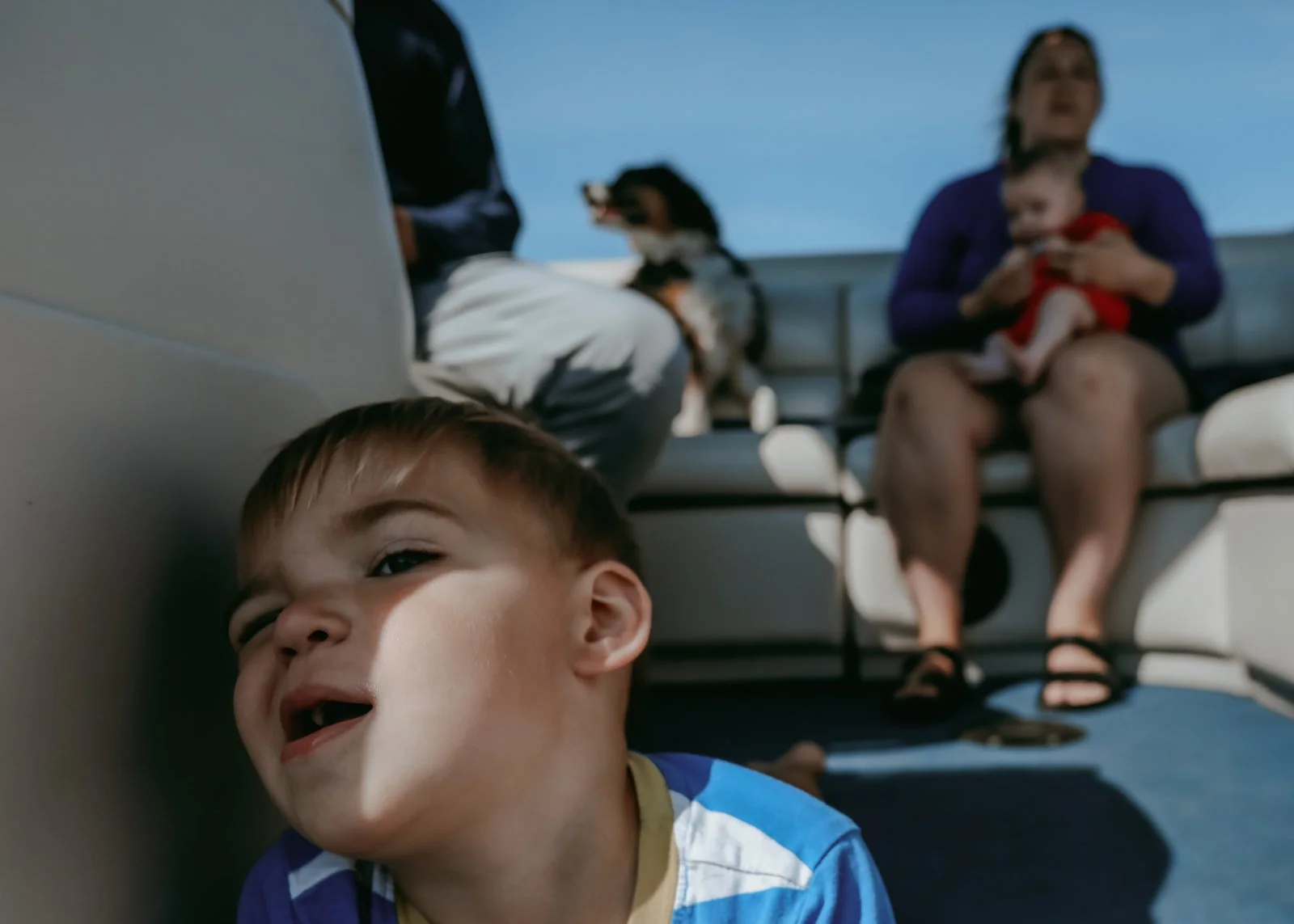 A young boy leaning against a boat wall, smiling with partial face shadow, with people in the background sitting on the boat's seats, and a woman holding a baby.