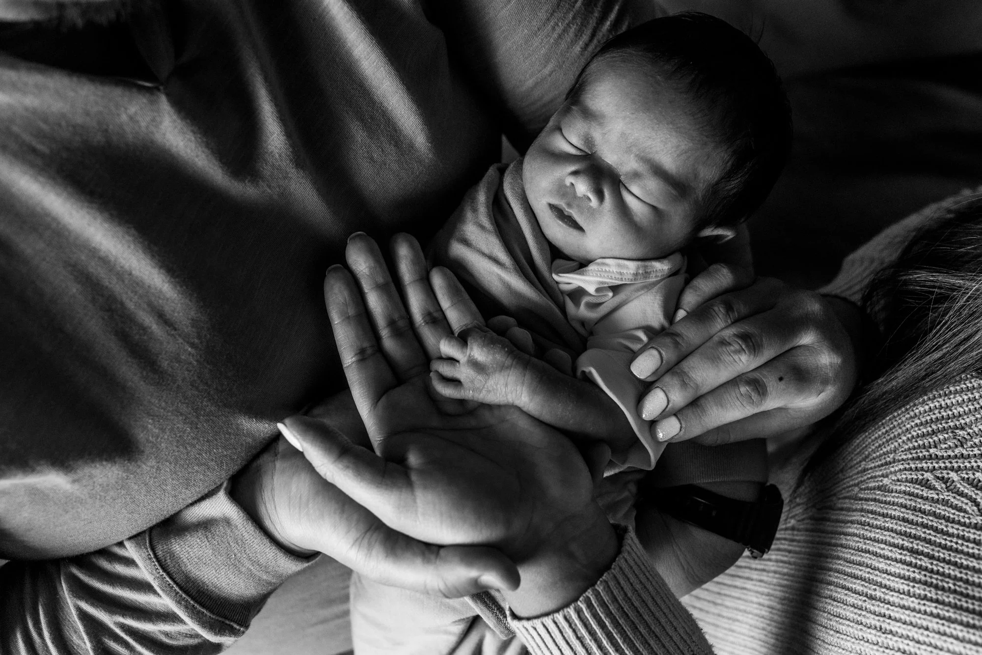 A group of adult hands gently holding a crying infant with closed eyes, in a close-up black and white photo.