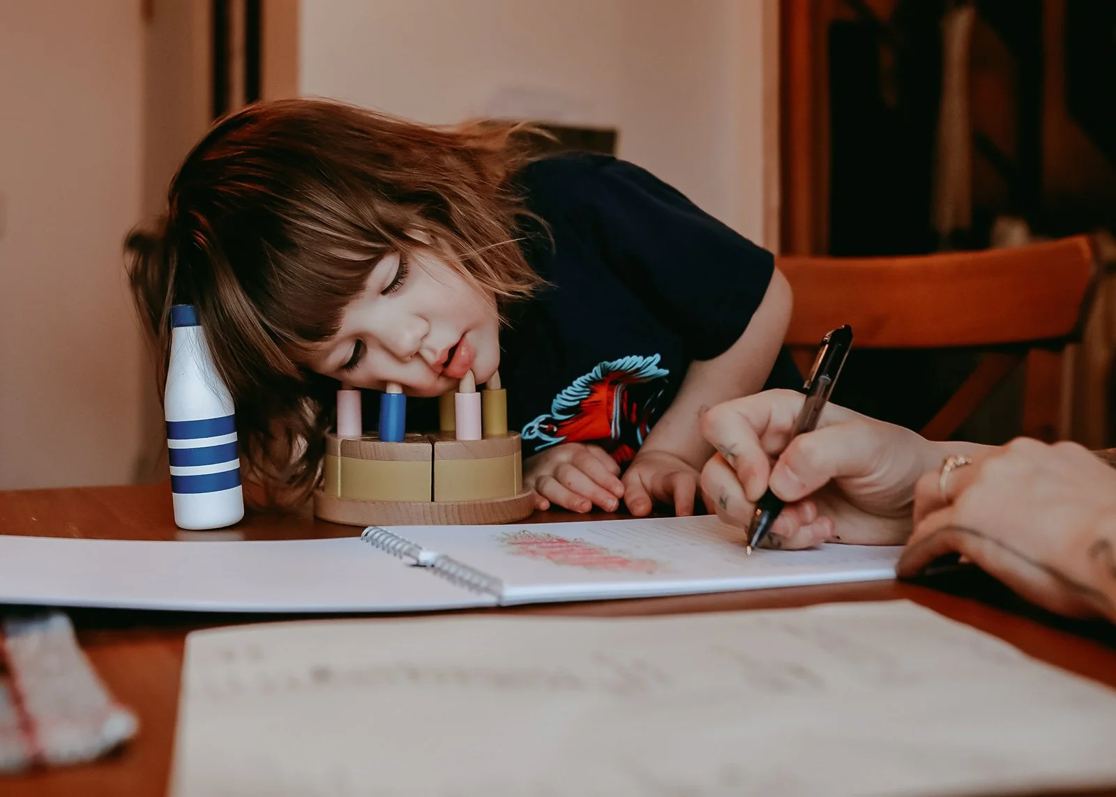 A young girl with short hair resting her head on a table, with her lips on a colorful wooden toy, while someone writes in a notebook nearby.