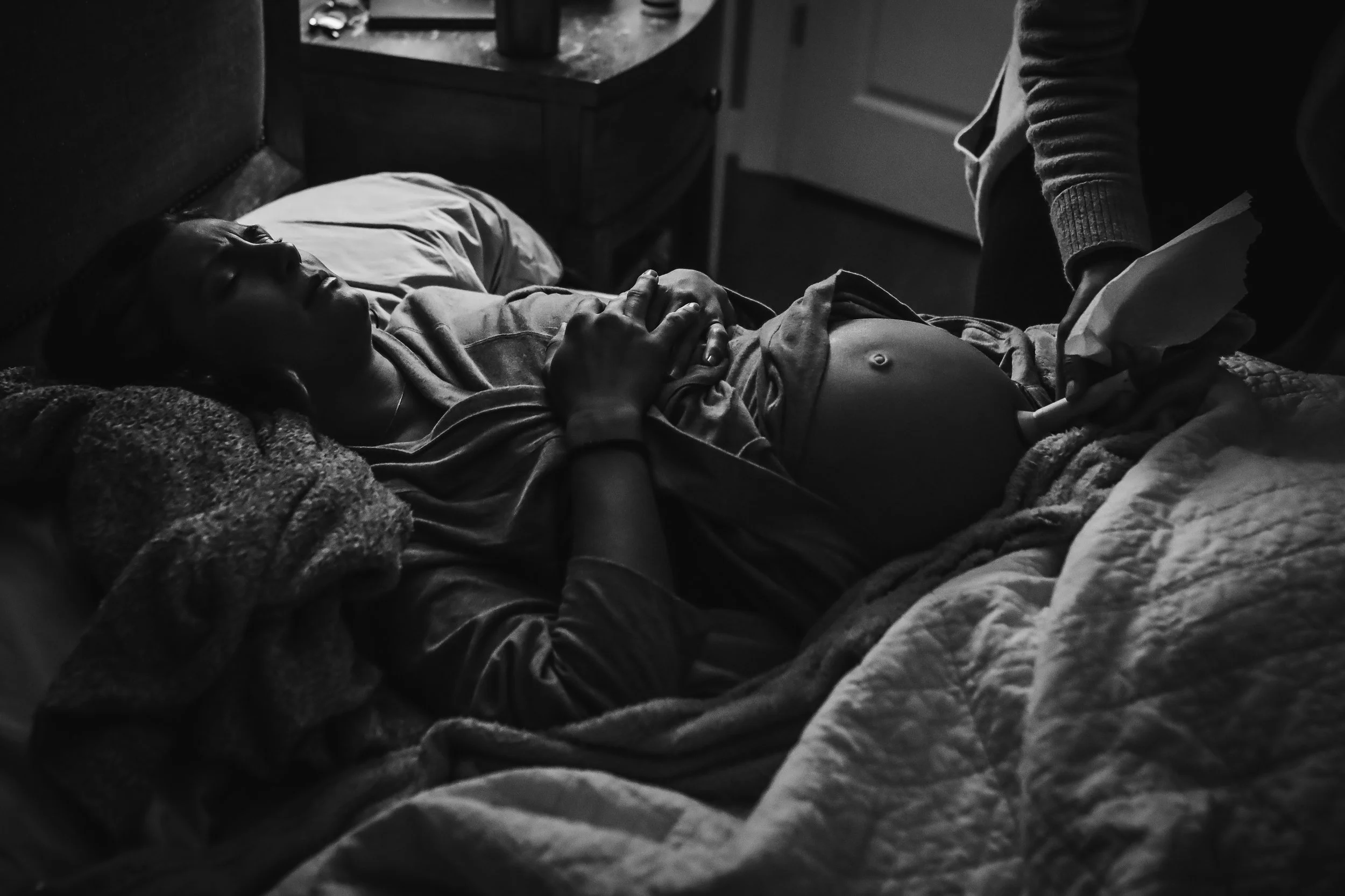 A woman lying in bed with her hand on her belly, which is being examined with a Doppler device held by a person standing nearby, in a dimly lit room.