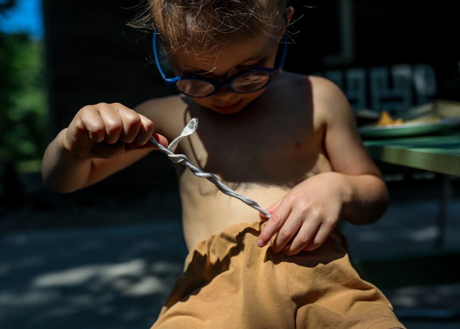 A young child with glasses, shirtless, outdoors during the daytime, holding and inspecting a spiral-shaped object.