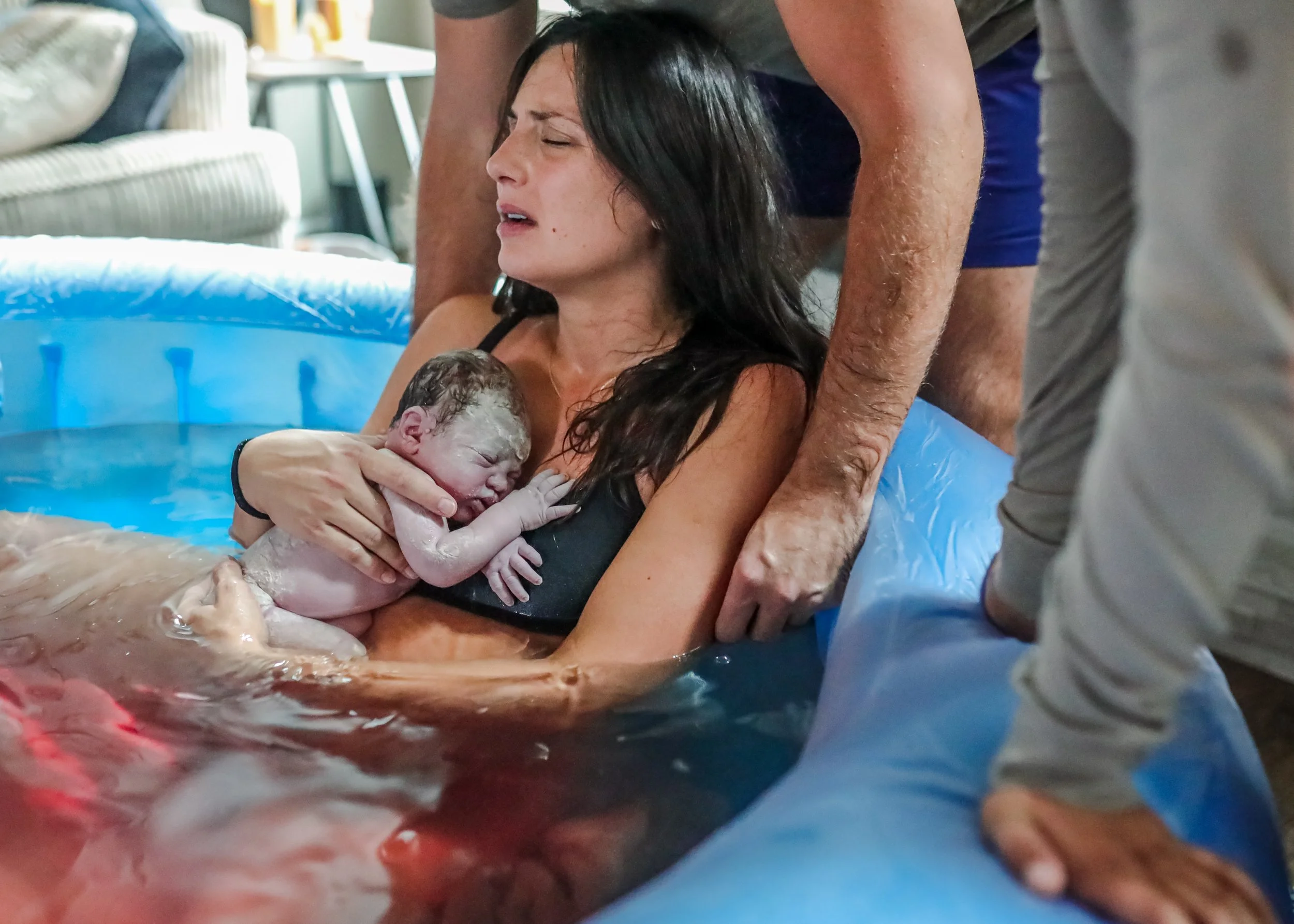 A woman giving birth in an inflatable birthing pool with a woman and a man assisting, as she holds her newborn baby.