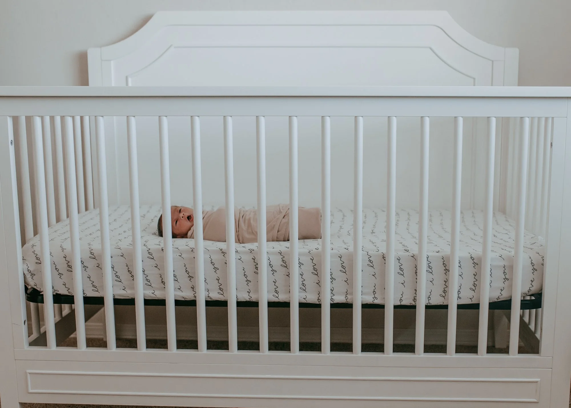 A baby lying in a white crib with a mattress that has black handwritten text, and the cribs bars are white, in a neutral-colored bedroom.
