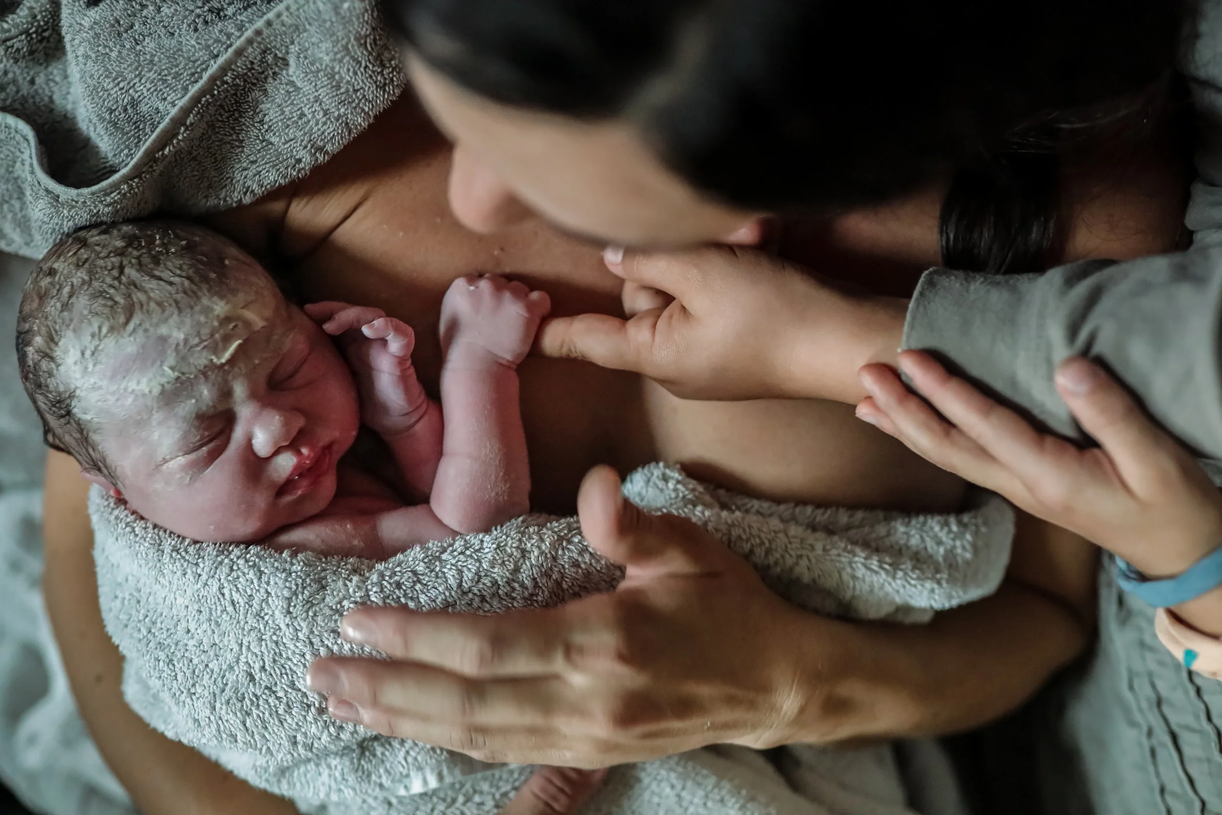 Newborn baby wrapped in a towel, being held and loved by a woman with long dark hair, while another person's hand gently touches the baby's foot.