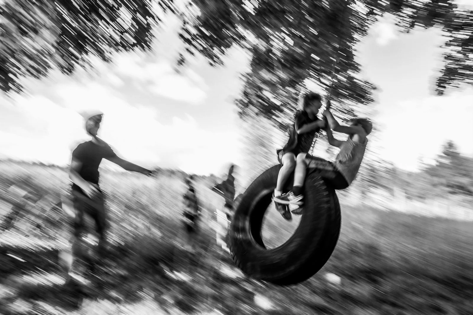 Two children playing on a tire swing hanging from a tree, with one child sitting on the tire and holding hands with another child who is standing nearby, while a person in the background watches.