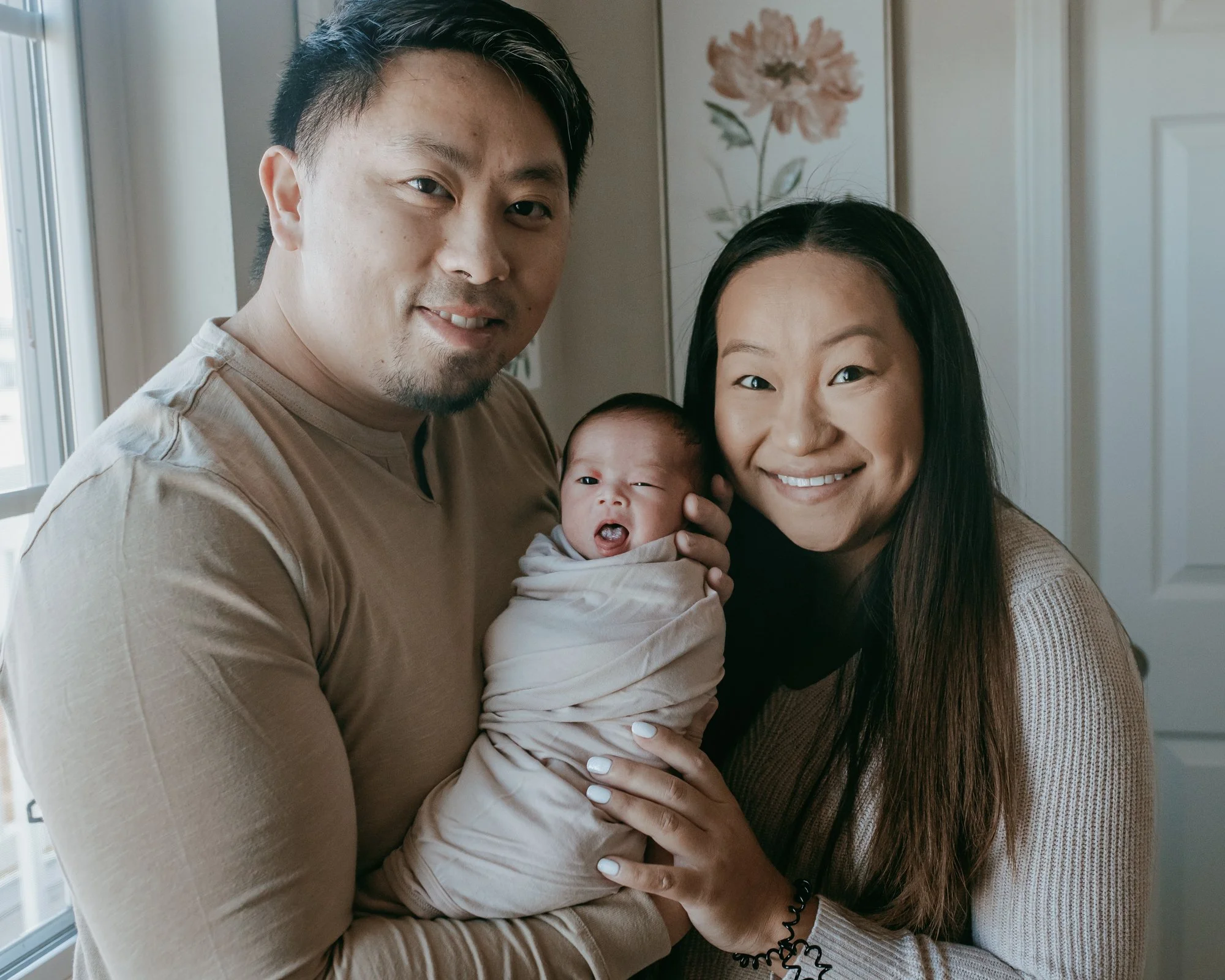 A mixed-ethnicity family of three with a man, woman, and newborn baby smiling indoors near a window.