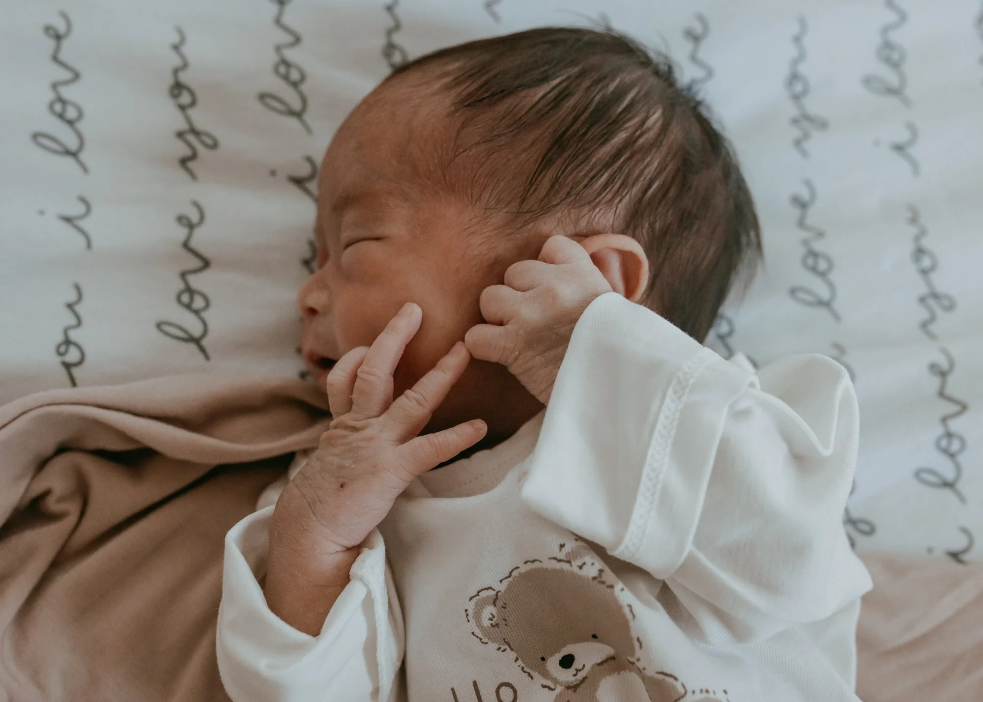A newborn baby sleeping on a pillow with writing, wearing a white onesie with a bear print, hand near face with fingers touching cheek and chin.