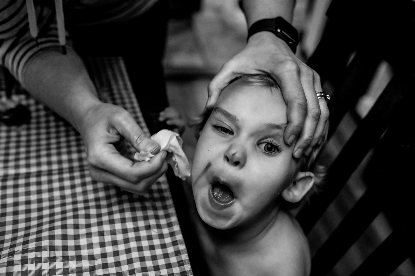 A young boy with wide eyes and mouth open, having his face painted by a person whose hands are visible.