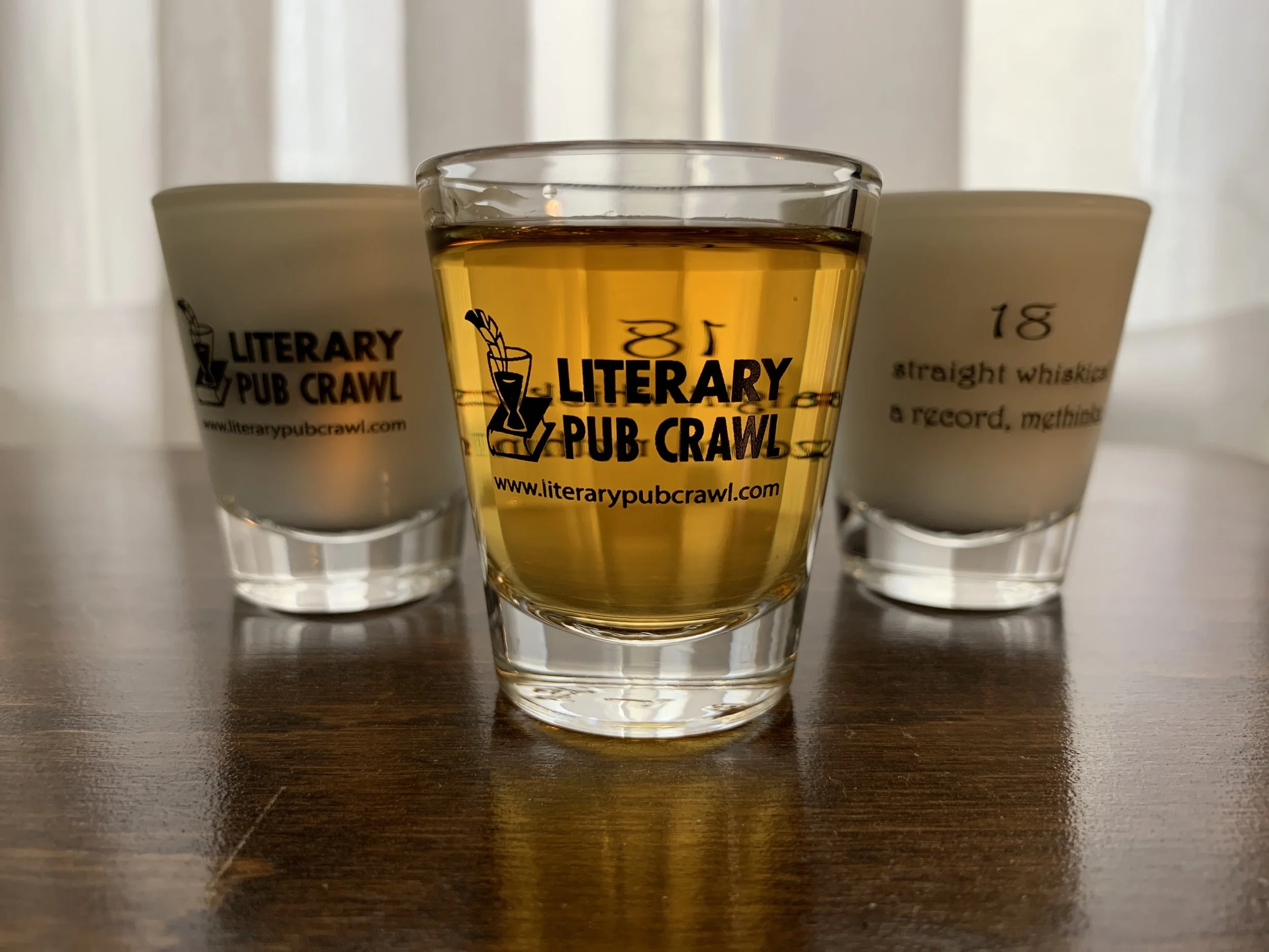 Three glasses on a wooden table with 'Literary Pub Crawl' logo and website. The center glass contains a golden liquid, and the two glasses in the background appear to hold white liquid, with writing visible on one.