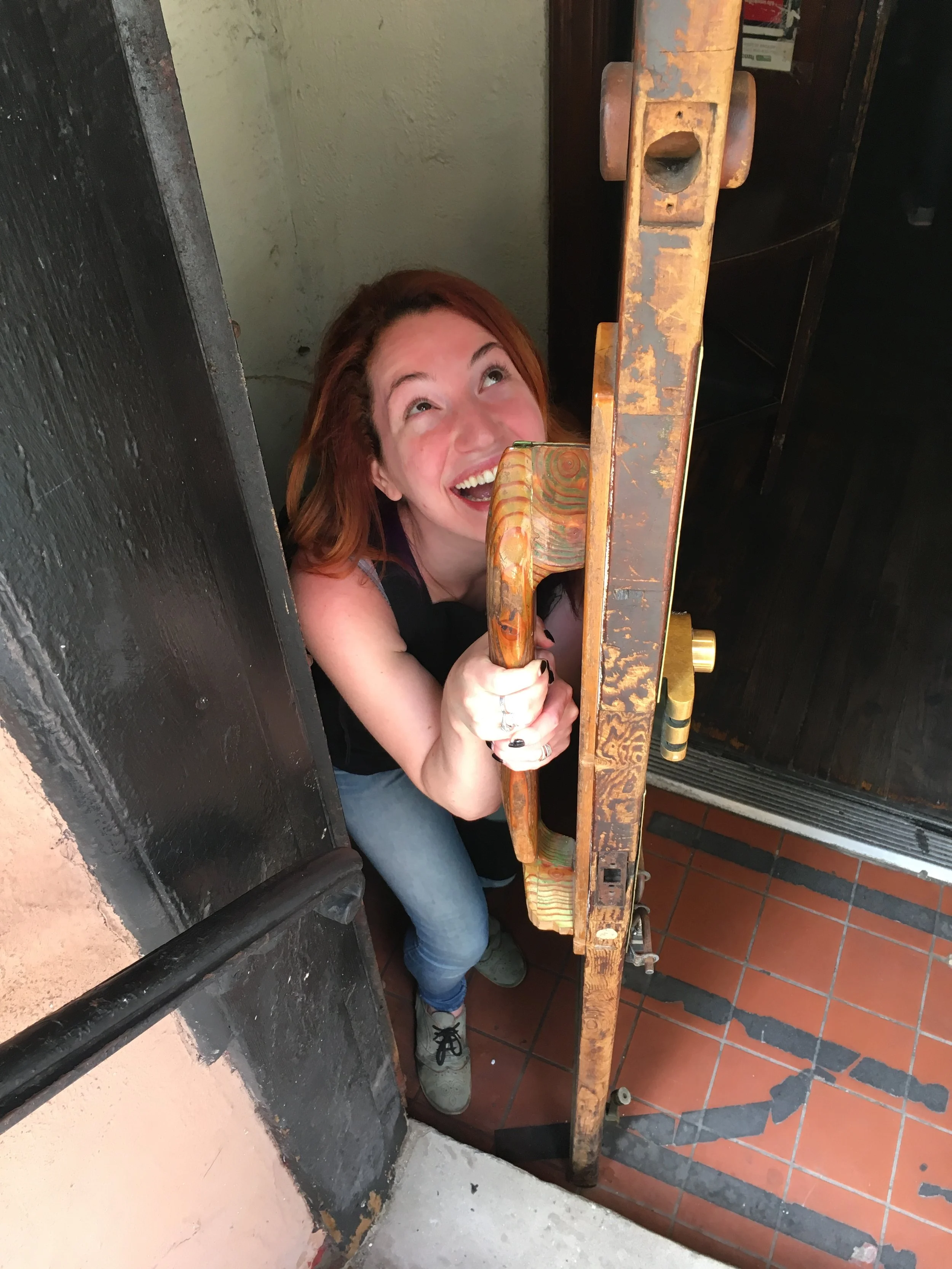 A woman with red hair smiling while peeking through a partially open door, holding the door handle with both hands.