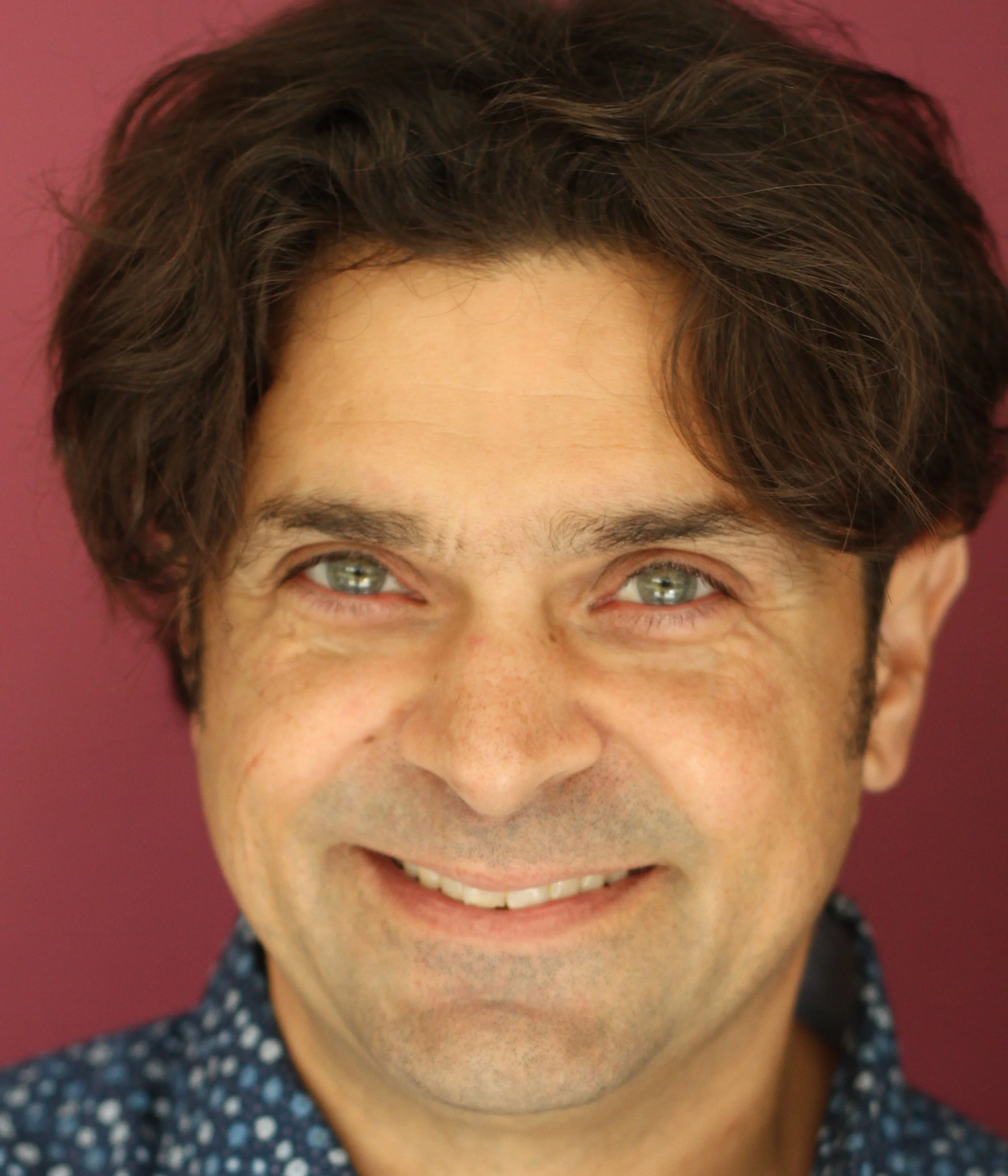 Close-up of a smiling man with brown hair and light-colored eyes, wearing a dark blue shirt with small white patterns, against a pink background.