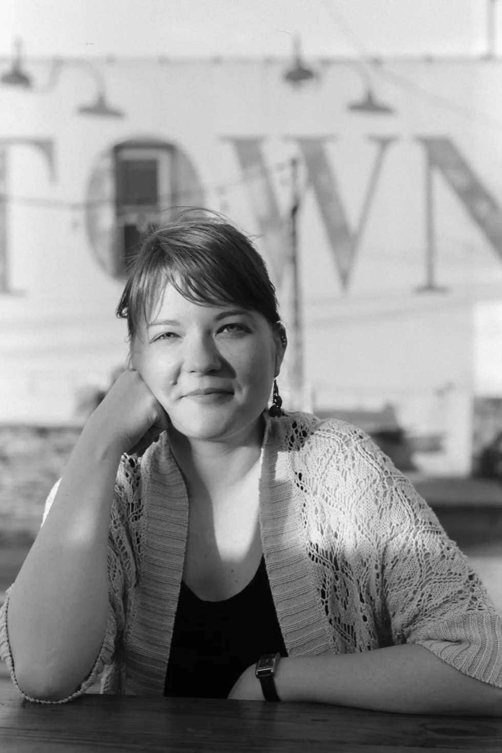 A woman sitting at a table with her head resting on her hand, wearing a lace sweater and earrings, with a partially visible sign in the background.