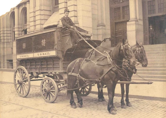 A historical black and white photo of a person riding in a U.S. Mail horse-drawn carriage in front of a large building with steps.