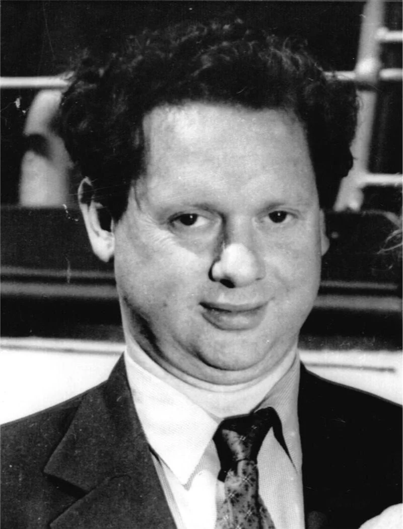 Black and white photo of a man with curly dark hair, light skin, wearing a suit and tie, smiling slightly.