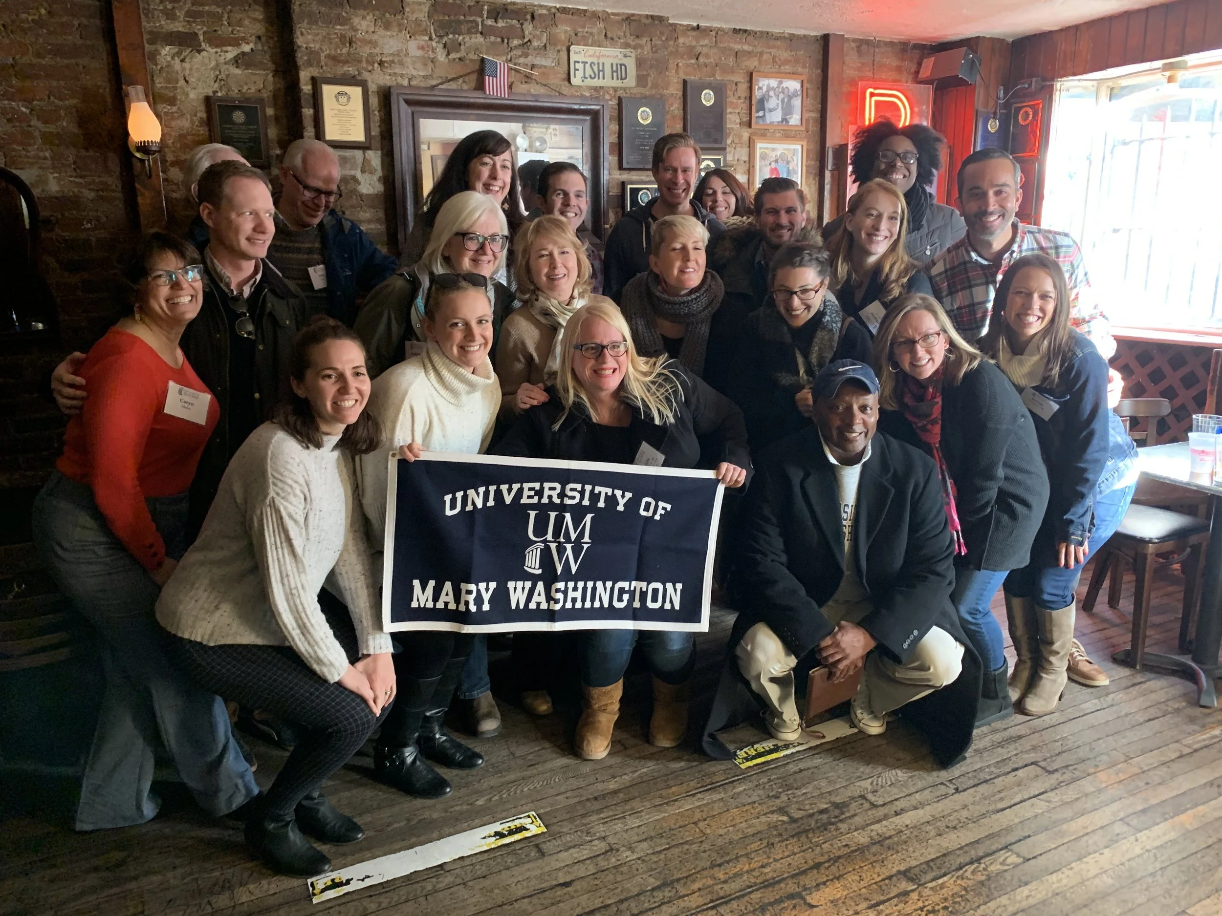 Group of people posing with a banner that reads "University of Mary Washington" inside a rustic restaurant or pub with brick walls and wooden floors.