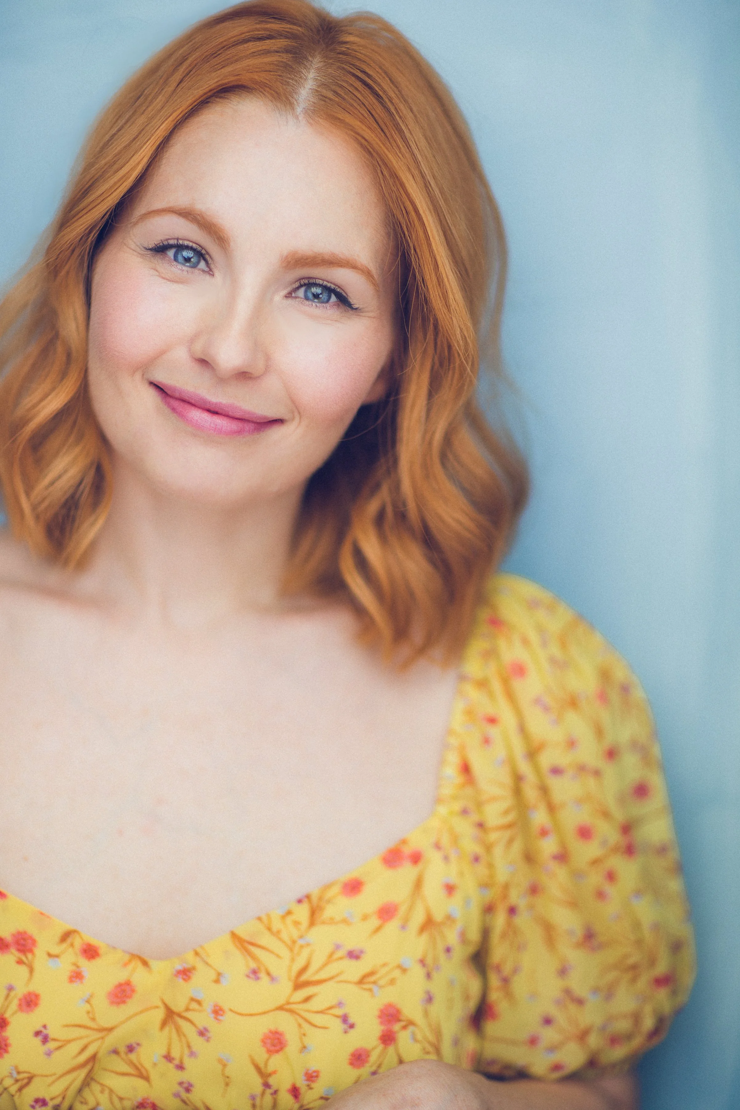 Portrait of a red-haired woman with blue eyes wearing a yellow floral top, smiling, against a light blue background.