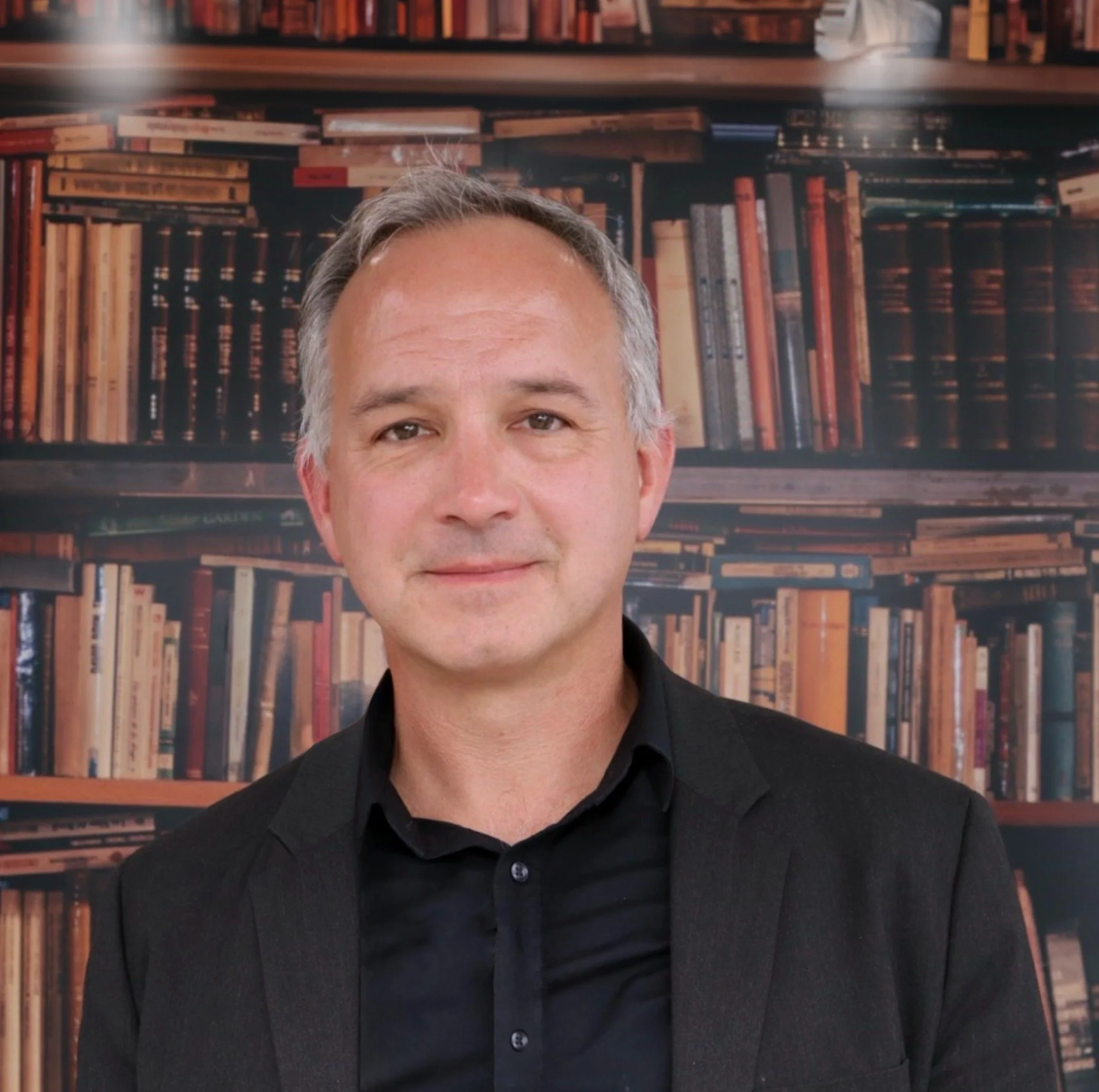 A man with gray hair and a light smile, wearing a black suit and shirt, standing in front of a background with a large bookshelf filled with books.
