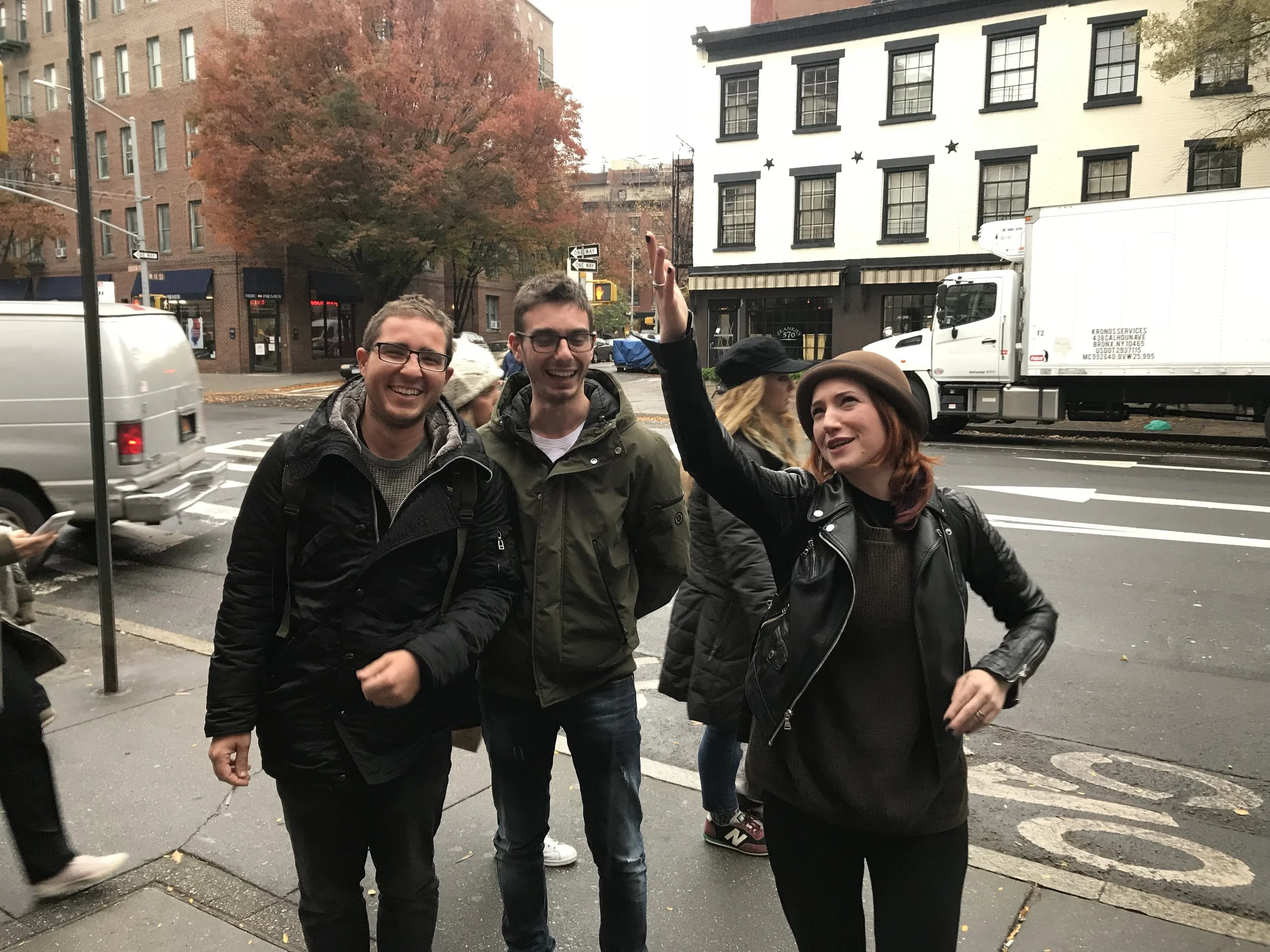 Three young people standing on a city sidewalk, smiling and waving. Two men on the left wearing jackets, a woman on the right in a black leather jacket and hat, with an autumn tree and buildings in the background.