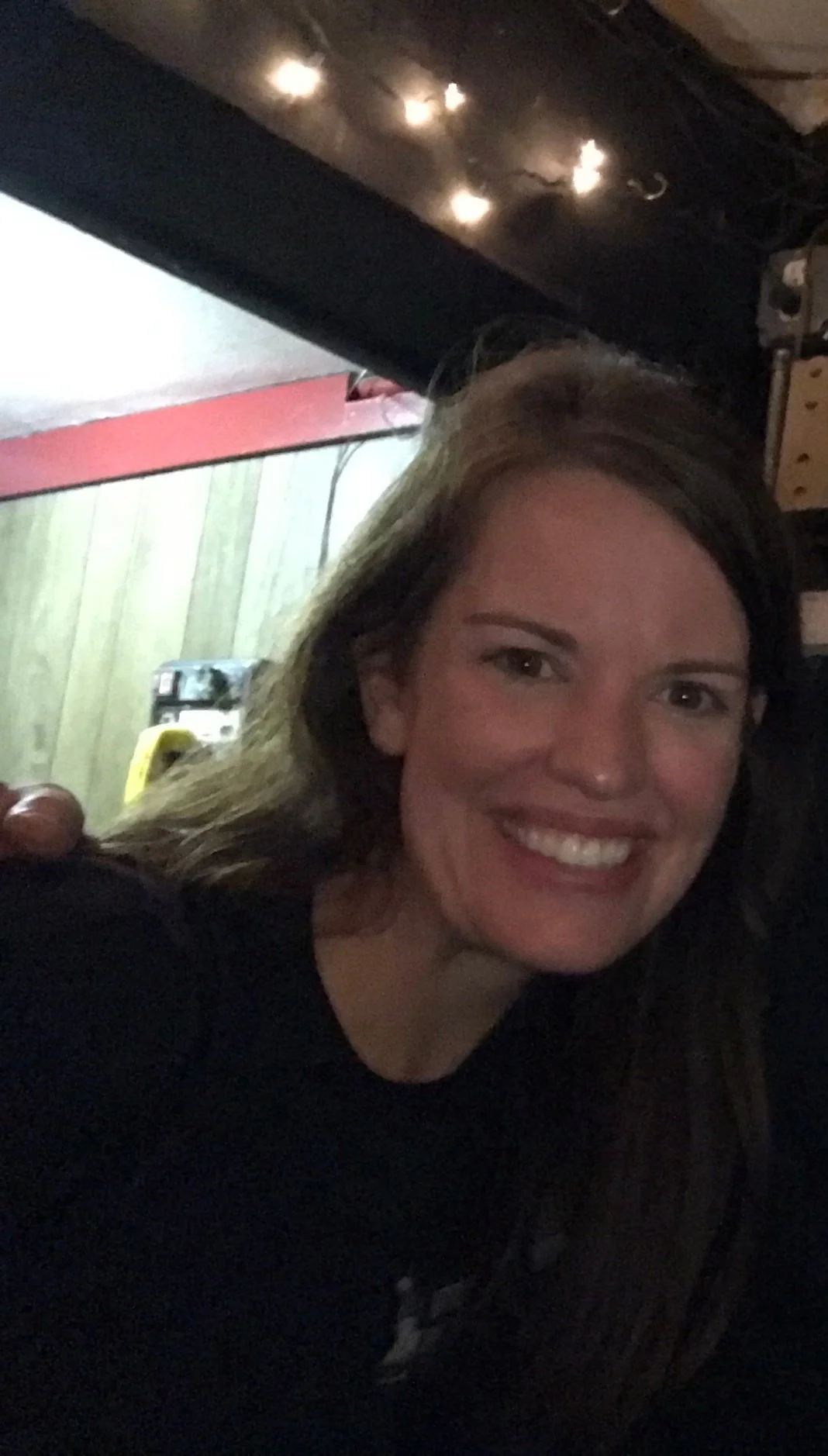 A woman with long brown hair smiling in a dimly lit room underneath a table or counter with string lights hanging above.